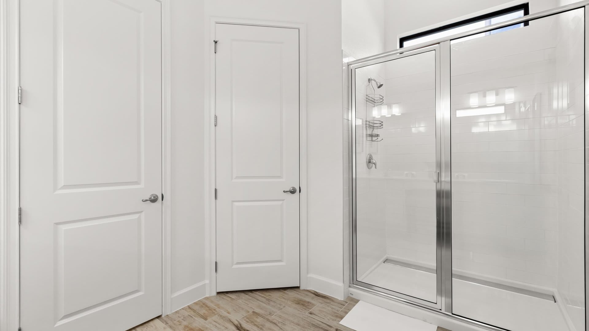 Modern glass-enclosed shower with white subway tile walls, chrome fixtures, and a built-in shelf. Two white panel doors are visible next to the shower, set against a wood-look tile floor.