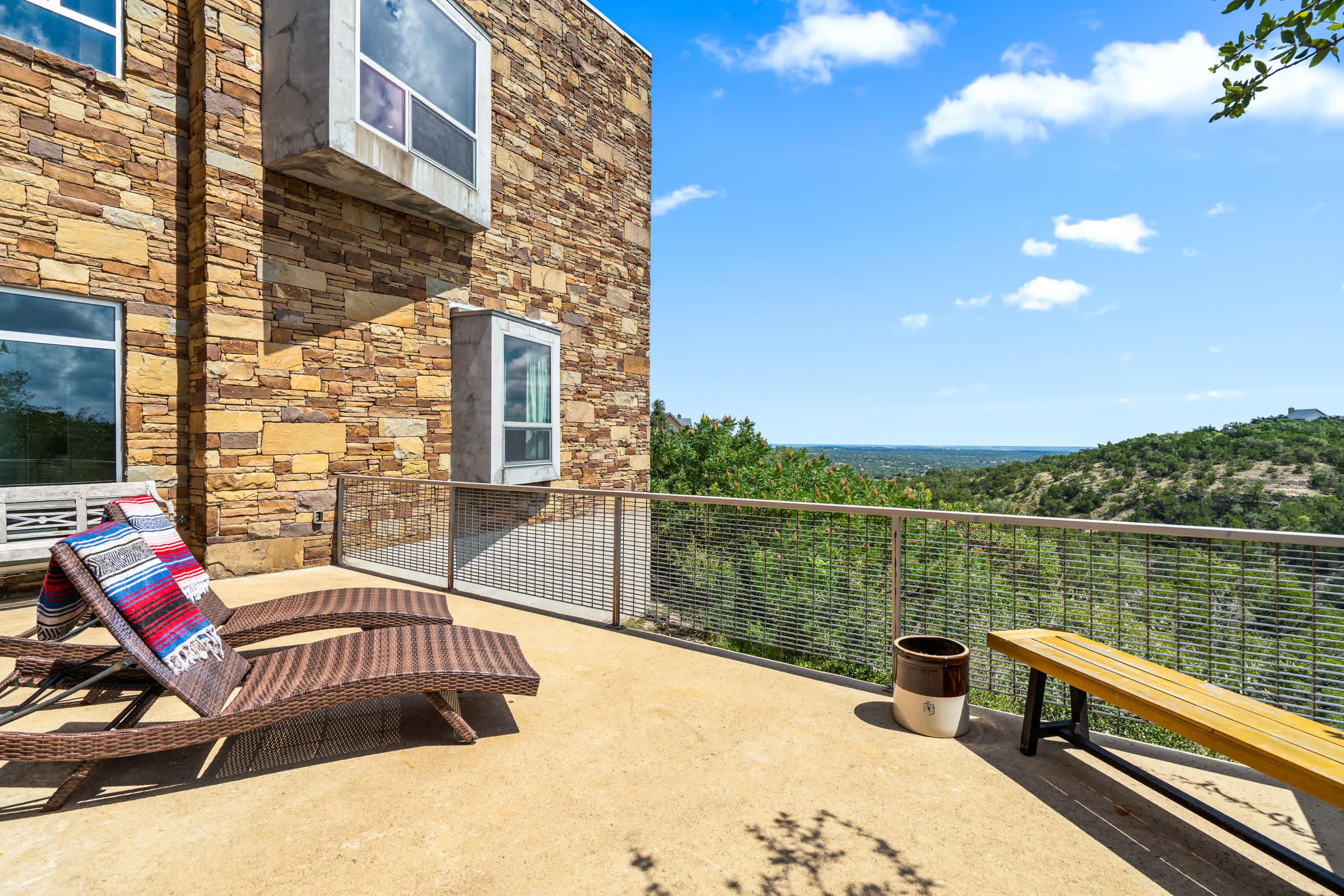 A spacious outdoor deck with two lounge chairs and a wooden bench offers breathtaking views of a sprawling valley and distant horizon. The modern house exterior features stacked stone cladding and large windows.