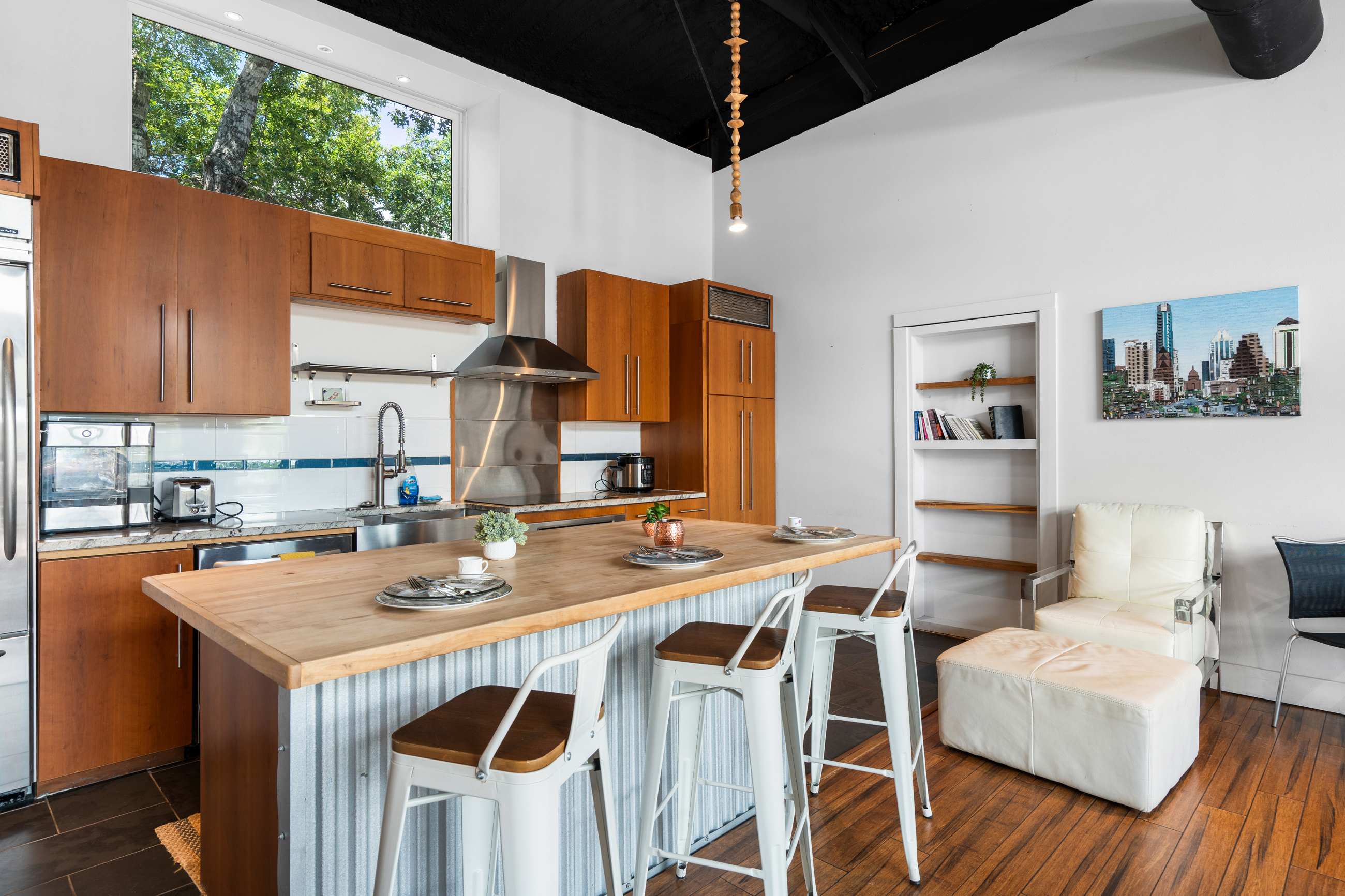 The kitchen showcases light wood cabinets, stainless steel appliances, and a large island with a light wood countertop and corrugated metal siding. A large window above the sink offers a view of trees, and a white bookshelf is visible in the background.