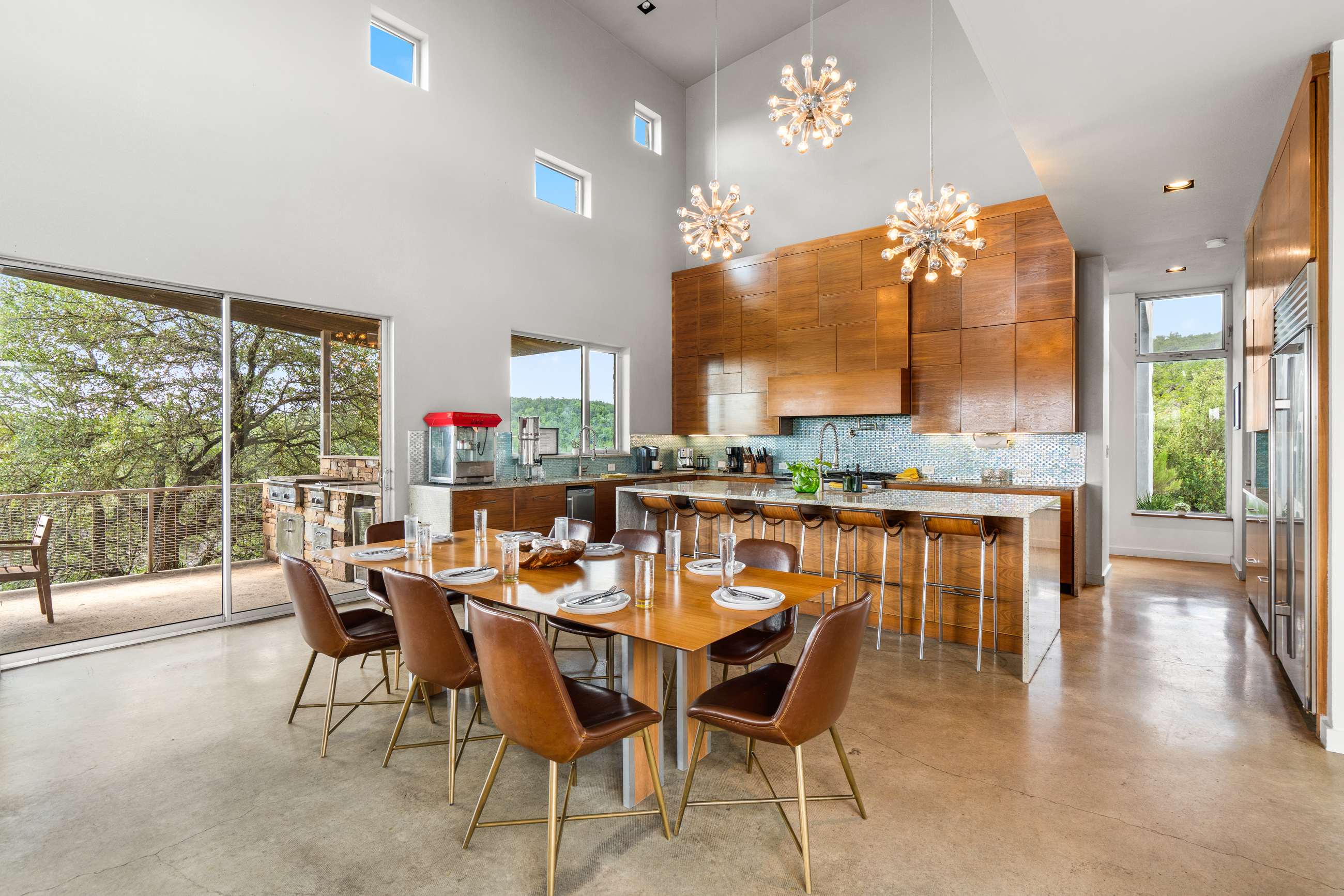 A modern open-concept kitchen and dining area featuring a long wooden dining table with brown leather chairs, a large kitchen island with bar stools, and high ceilings with multiple windows and starburst chandeliers. Large sliding glass doors open to an outdoor area.