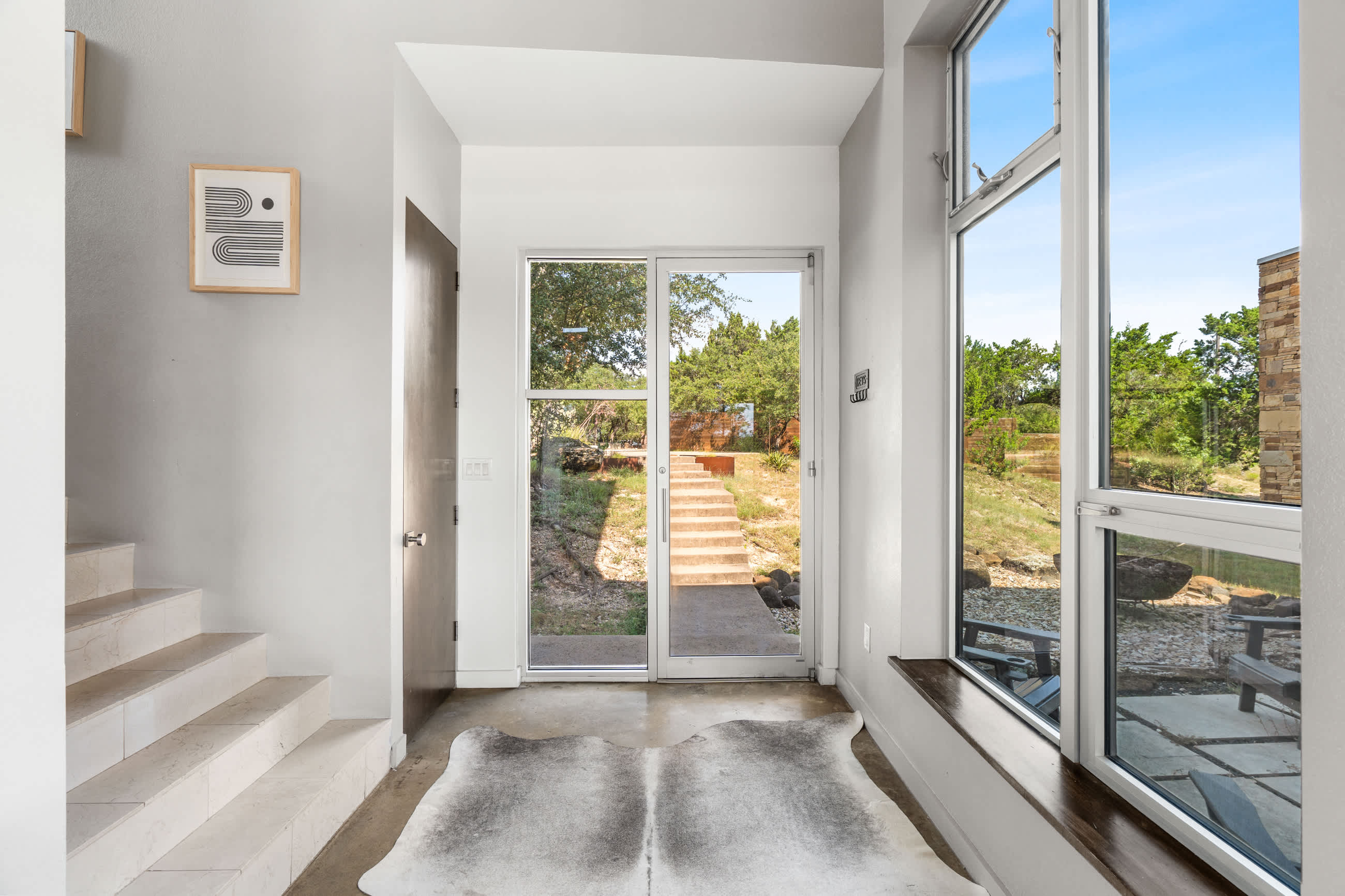 A minimalist foyer with light gray walls, polished concrete floors, and a cowhide rug, leading to a staircase and offering views of the outdoor area through large glass doors and windows.
