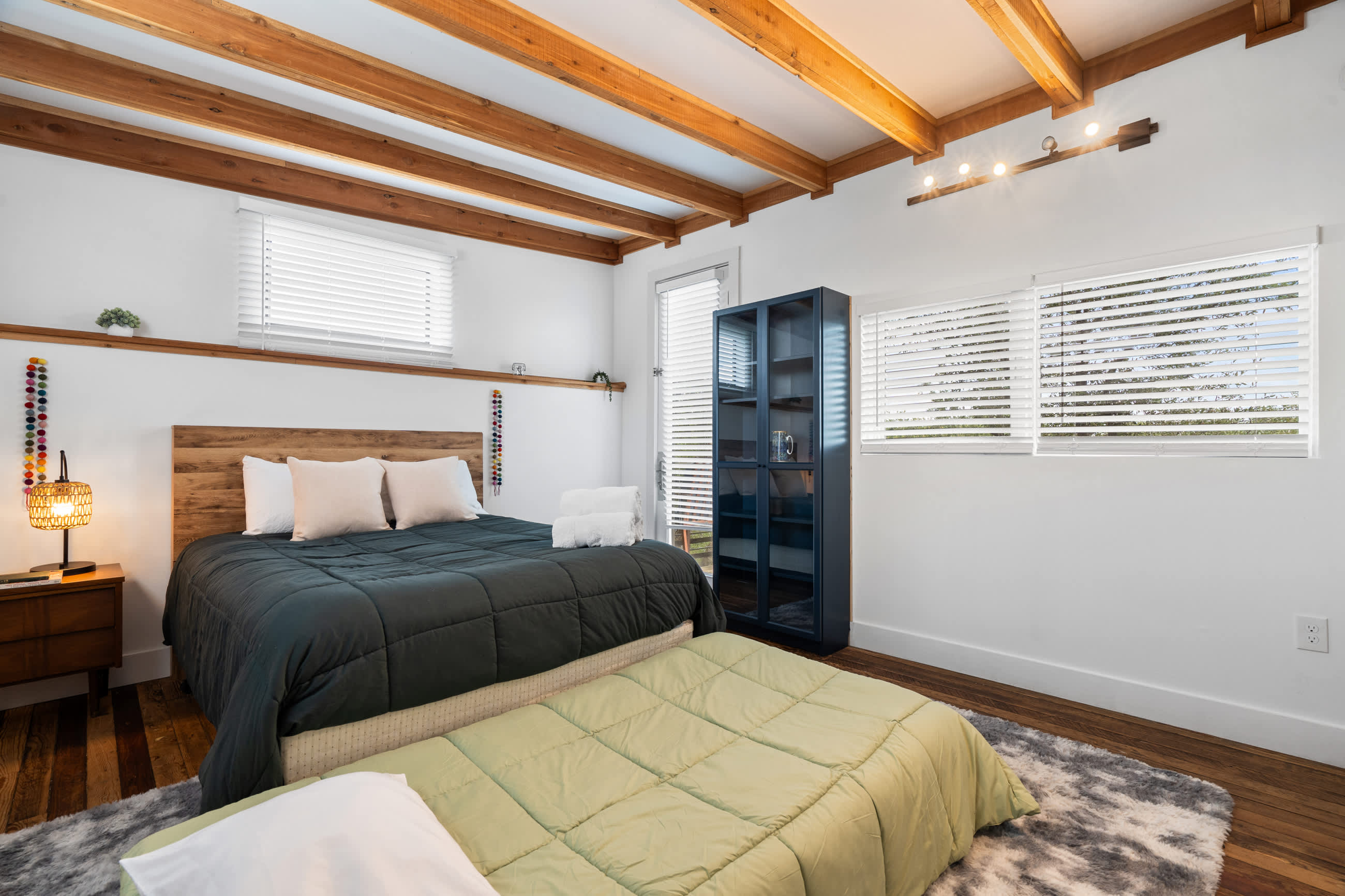 Wide shot of the bedroom, showcasing the queen bed with a wooden headboard, a nightstand with a lamp, and a dark blue shelving unit. The room features white walls, exposed wooden ceiling beams, hardwood floors, and multiple windows with blinds.