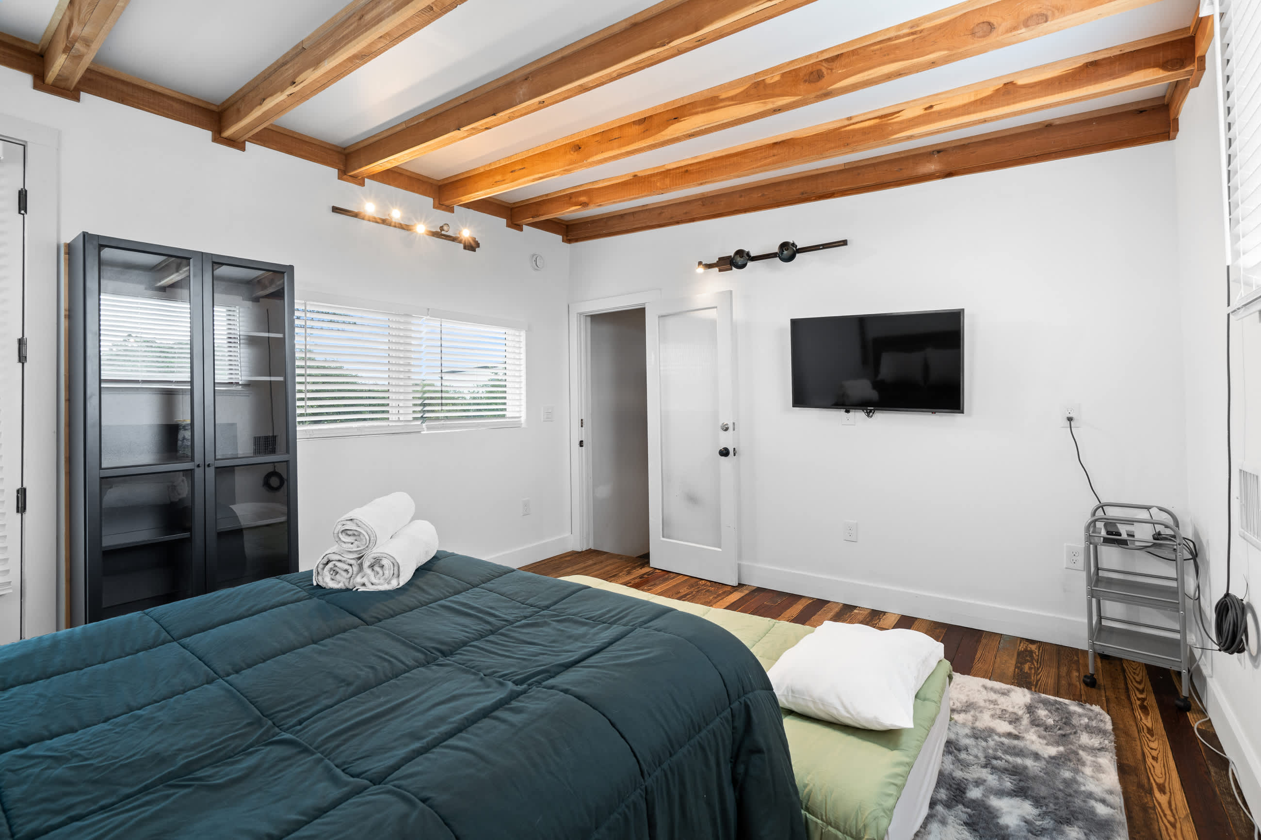 Another perspective of the bedroom, highlighting a dark metal and glass cabinet, the wall-mounted TV, and the bed with white rolled towels. The room maintains its bright white walls, exposed wooden beams, and hardwood floors.