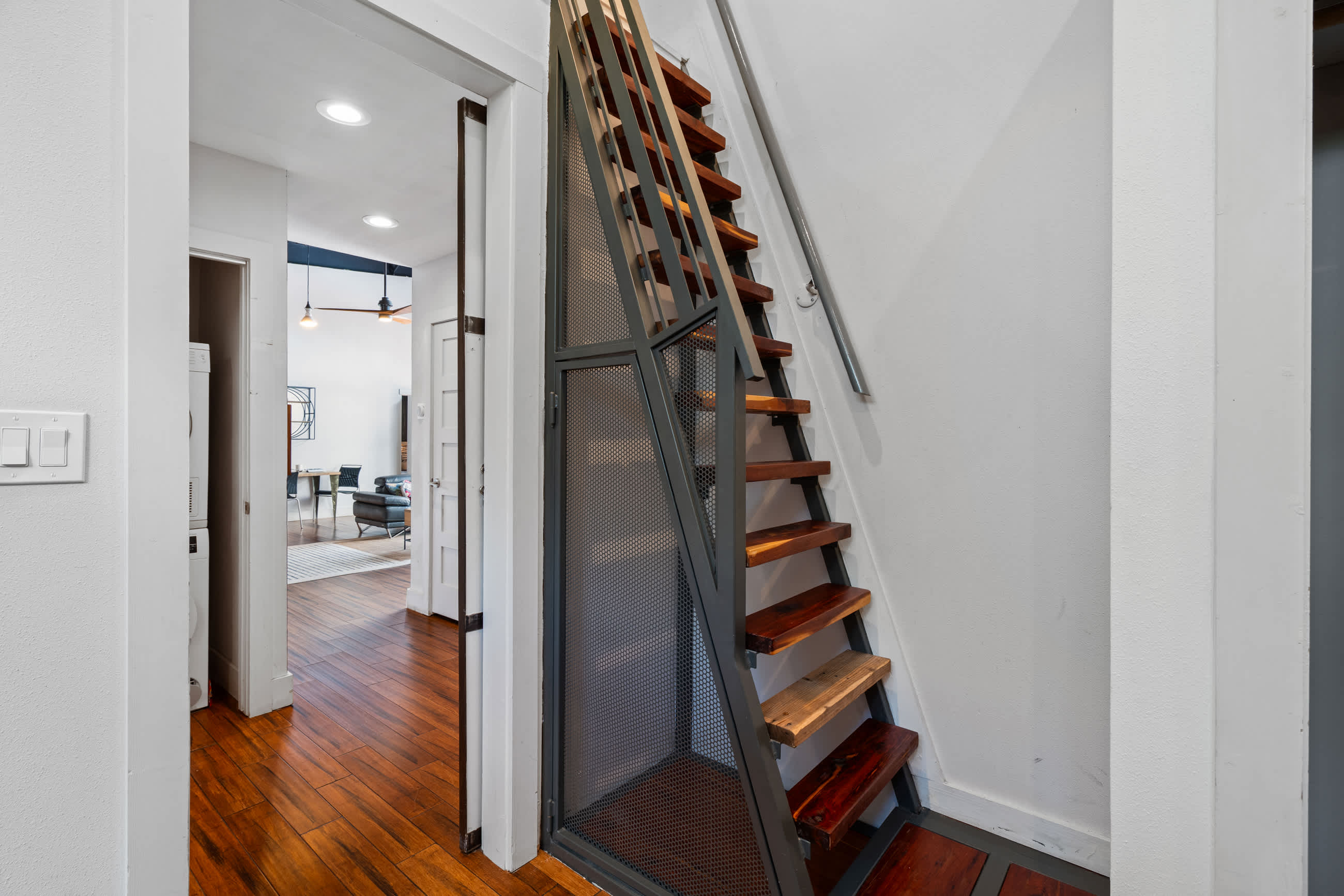 A modern industrial-style staircase with wooden treads and metal railings leads upstairs, adjacent to a doorway opening into another room. Dark hardwood floors extend through the hallway.