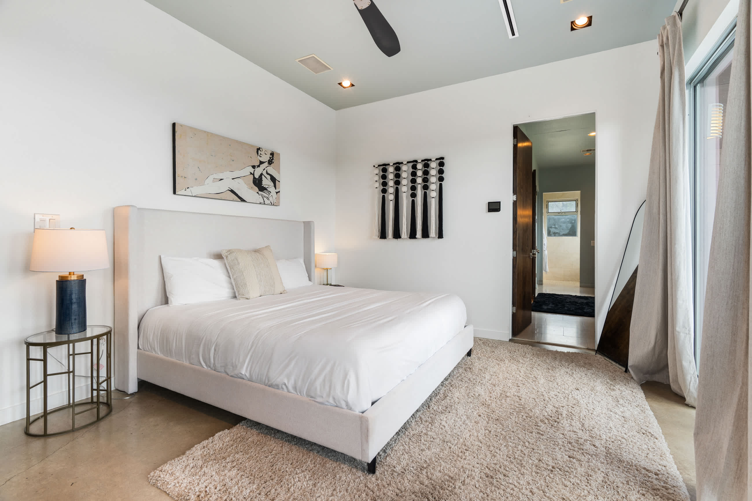 A serene bedroom featuring a white upholstered king-size bed with crisp white bedding and a decorative pillow. A shaggy area rug covers the concrete floor, and a modern side table with a lamp sits beside the bed. A doorway leads to an en-suite bathroom.