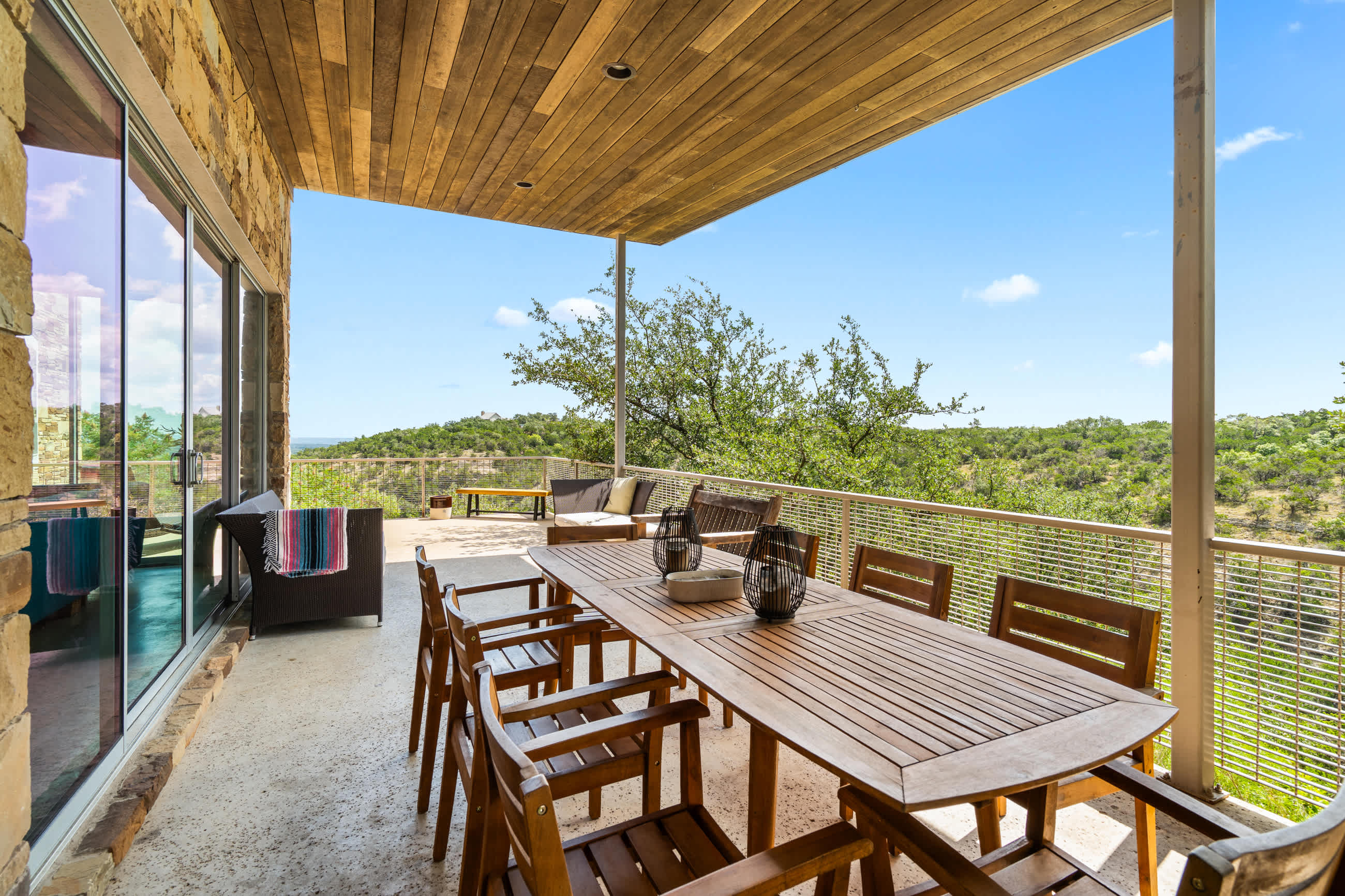 Spacious covered patio with a large wooden dining table and multiple chairs, along with additional outdoor seating, all overlooking a vast landscape of green hills. The ceiling is wood-paneled, and a glass sliding door leads into the interior.