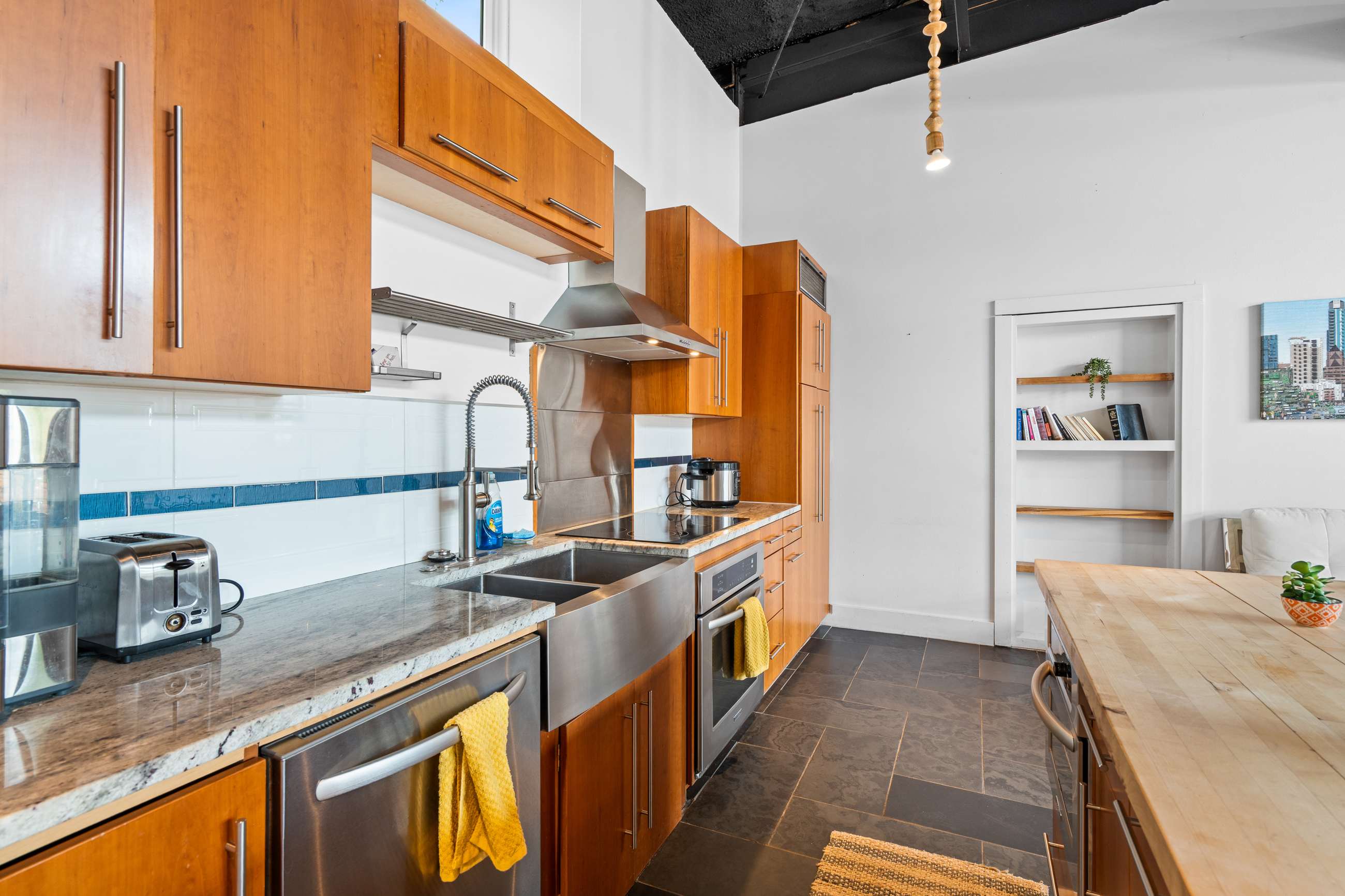 A close-up of the kitchen counter reveals granite countertops, a large stainless steel sink, and an induction cooktop. Light wood cabinets line the walls, and a white subway tile backsplash with a blue accent stripe adds a pop of color.