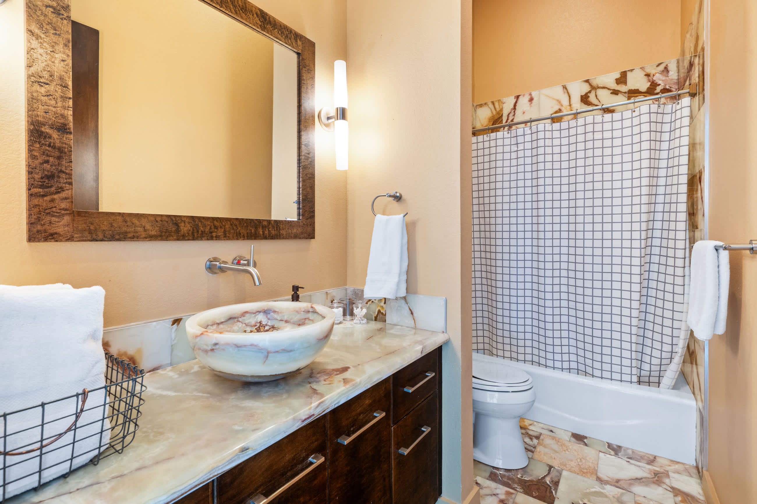 A warm-toned bathroom featuring a unique stone vessel sink on a marble-topped vanity, a large wooden-framed mirror, and a shower/tub combo with a grid-patterned curtain and marble-tiled walls.