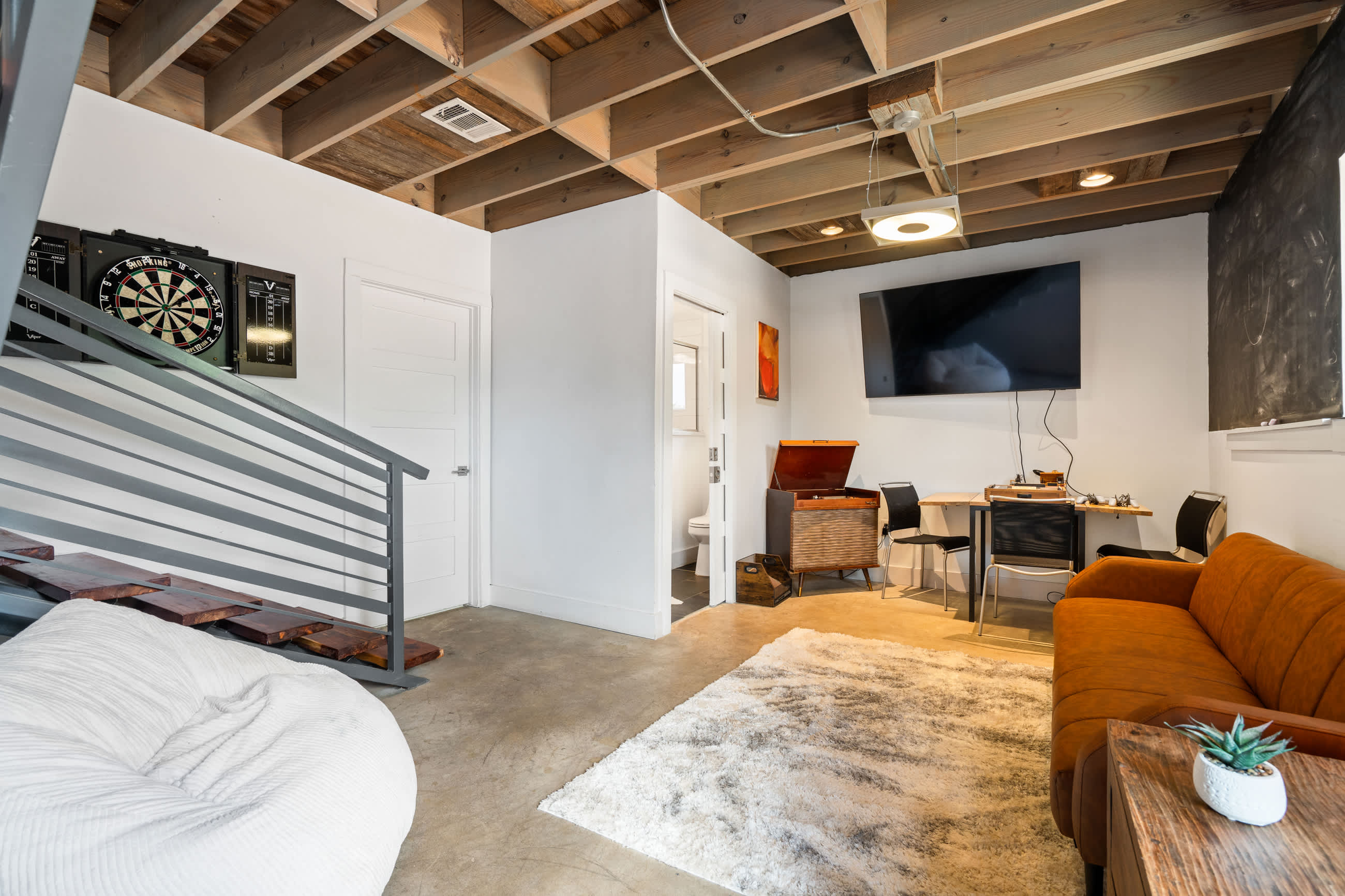 A recreation room features an exposed wooden beam ceiling, concrete floors, and a dartboard on the wall. A white bean bag chair is in the foreground, and a doorway leads to another room.