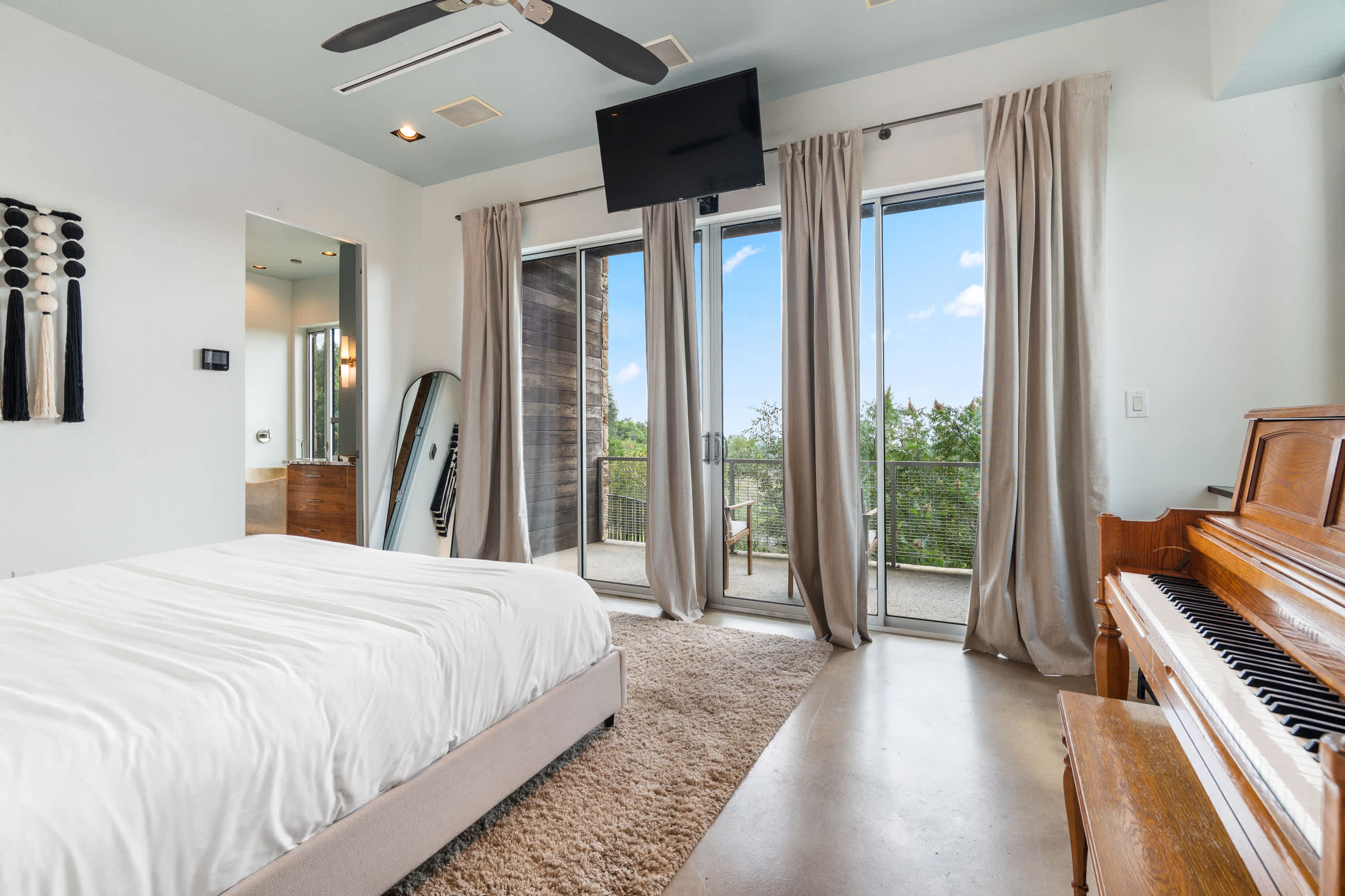 Modern bedroom with a white bed, shaggy rug, and large glass sliding doors leading to a balcony with outdoor seating and scenic views. A ceiling fan and wall-mounted TV are also visible.
