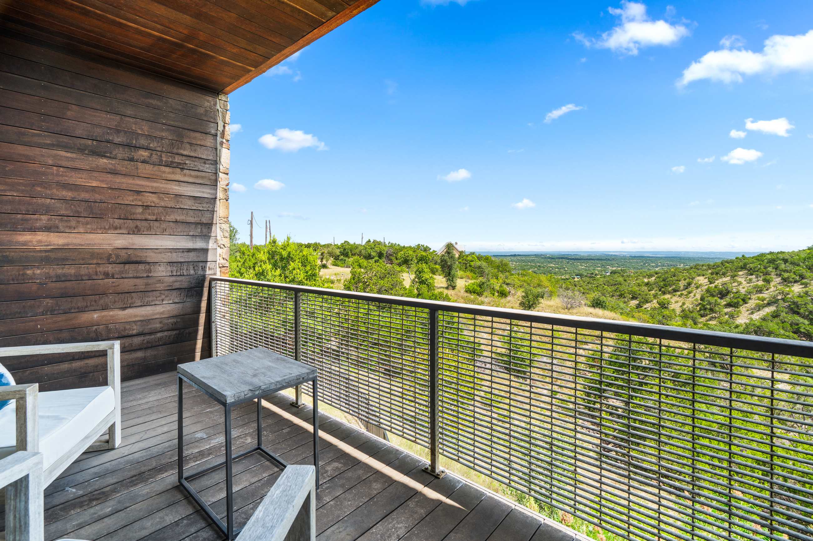 Balcony with a rich wooden slatted wall and wooden floor, featuring a small dark side table and a wooden chair with a white cushion, all set against a backdrop of expansive green hills and a bright blue sky.