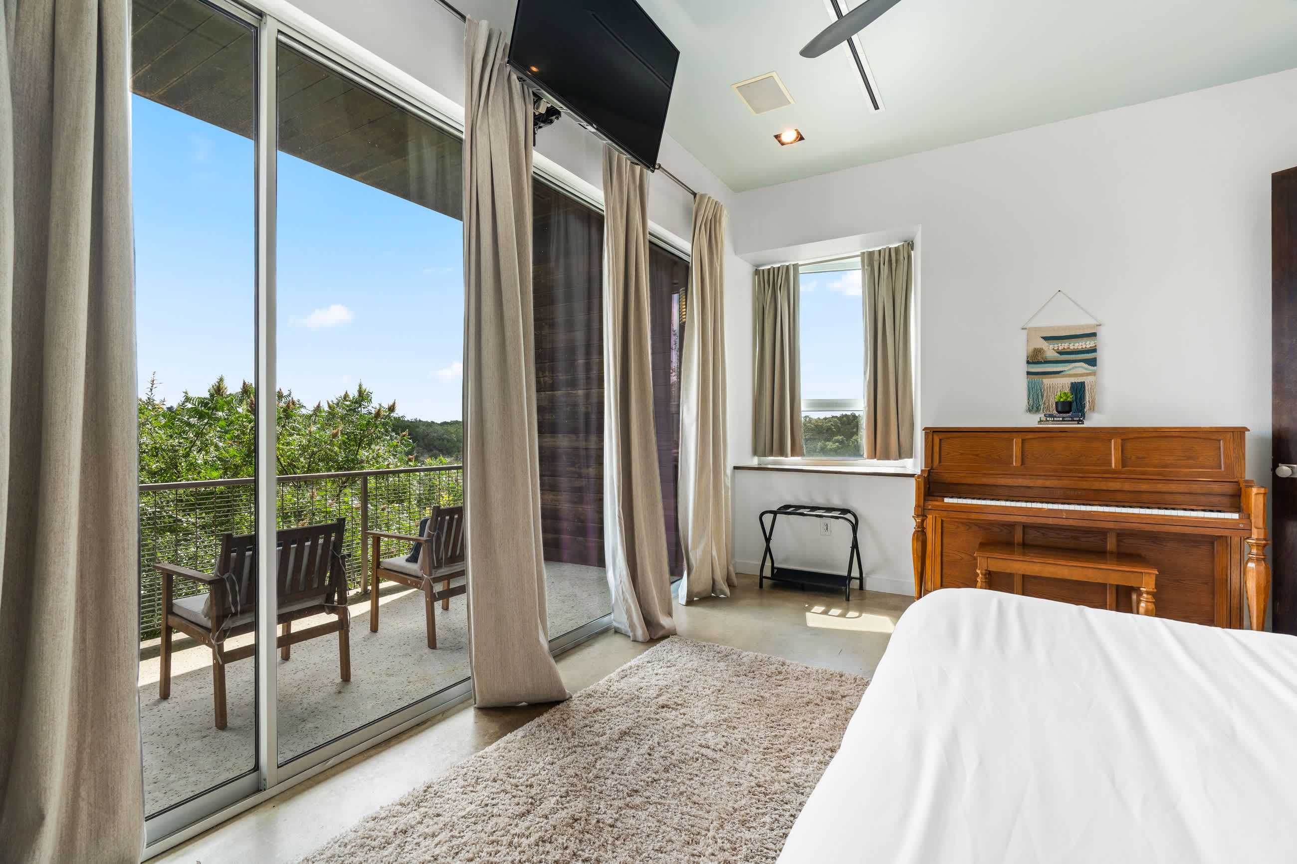 View of a spacious bedroom with a white bed, shaggy rug, and expansive glass sliding doors opening to a balcony with chairs and a panoramic view of the landscape. A wooden piano is visible on the right.