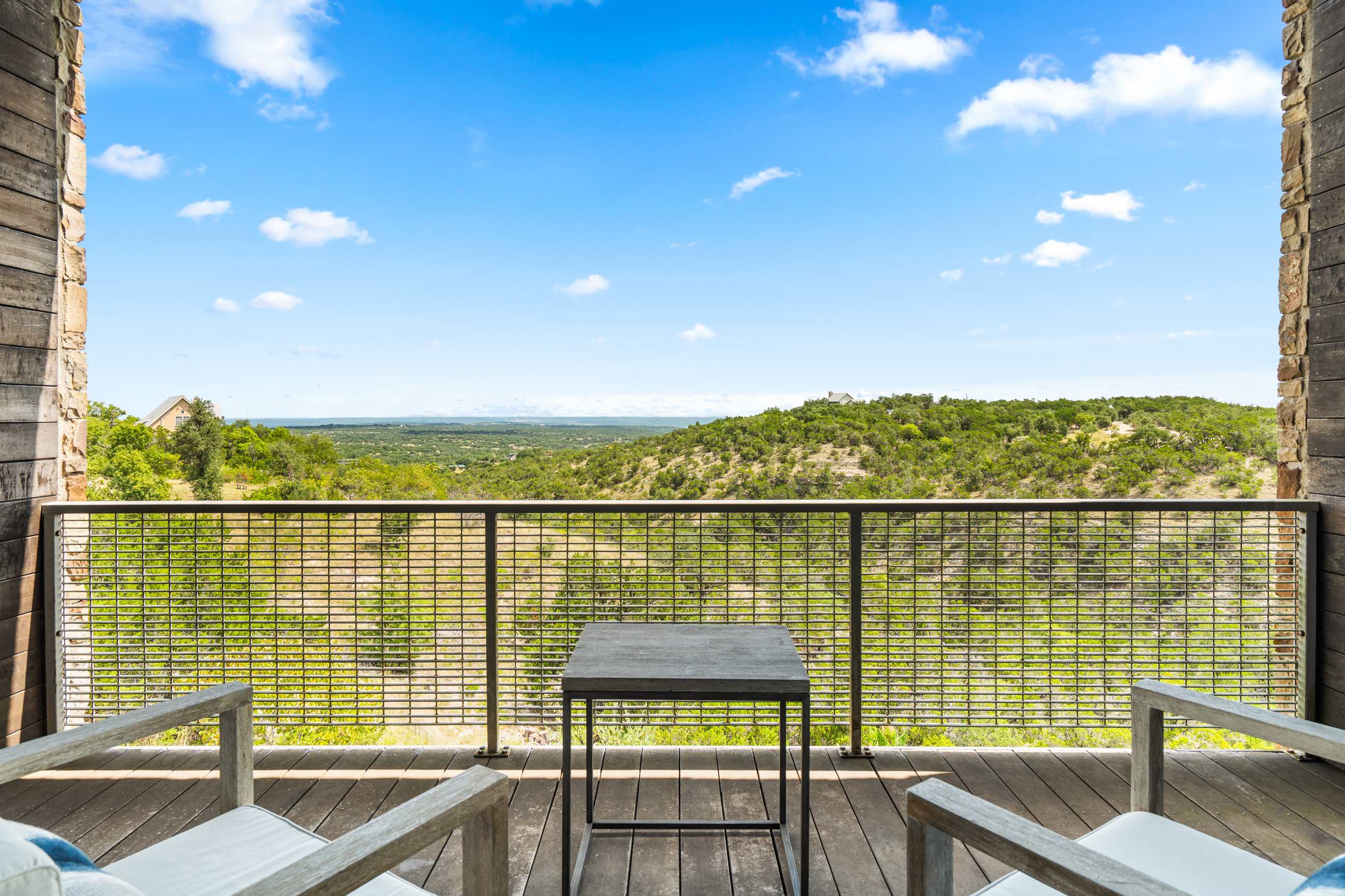 Balcony with a wooden floor and metal grid railing, featuring two wooden chairs with white cushions and a small dark side table, all facing a breathtaking view of distant green hills under a clear blue sky.