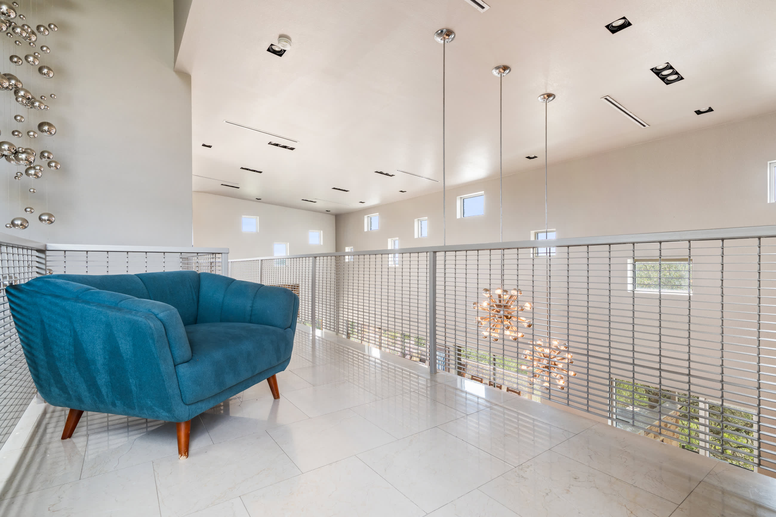Bright, open loft area with white tiled floors and a modern blue armchair, featuring a metal grid railing overlooking a lower level. Contemporary pendant lights hang from the high ceiling, and small windows line the upper walls.