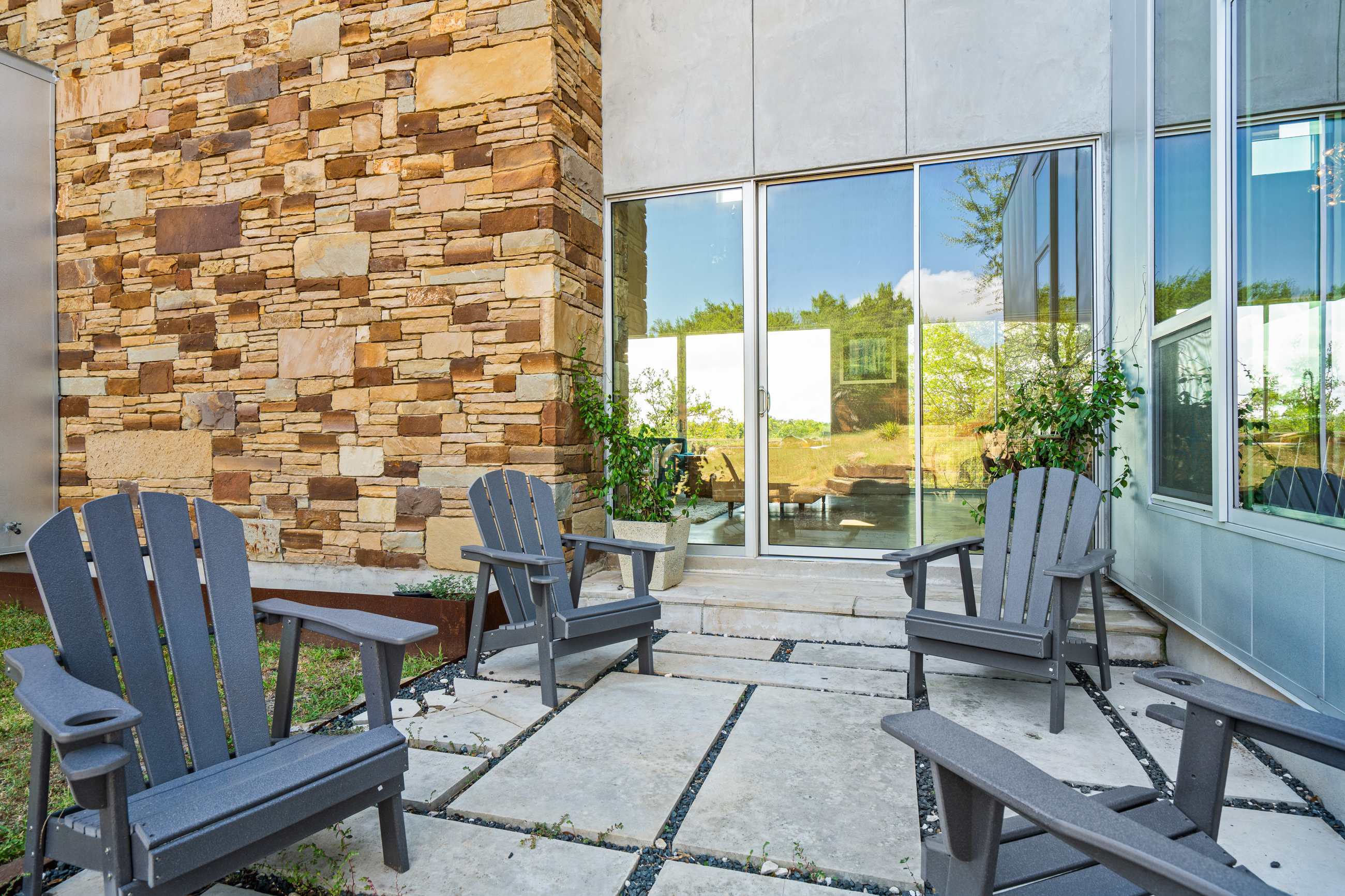 A modern patio features gray Adirondack chairs arranged on concrete pavers with dark gravel, set against a house with a stone facade and large glass sliding doors.
