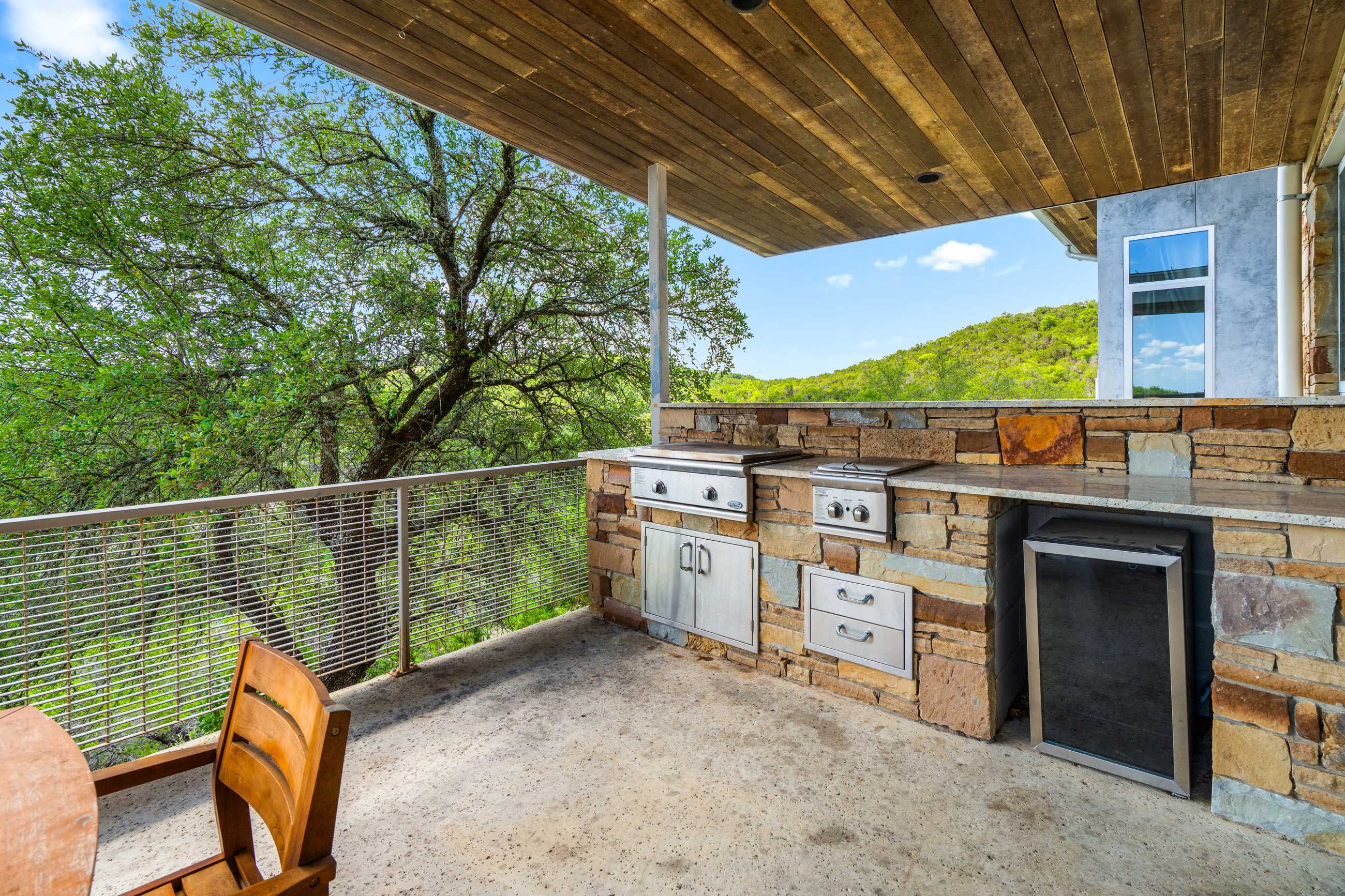 A modern outdoor kitchen, built into a stone wall, includes a stainless steel grill, side burner, and beverage fridge, with a wooden chair in the foreground. The patio overlooks a lush green landscape with a metal railing.