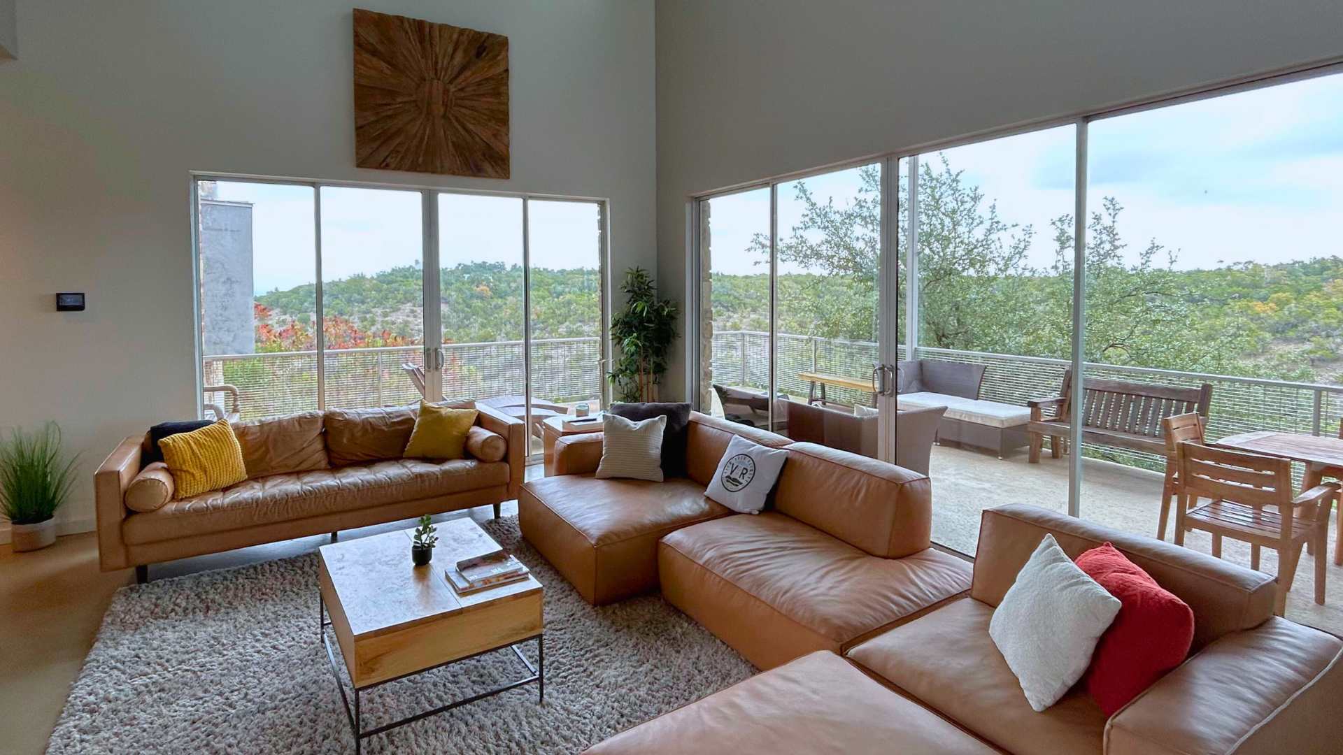 Bright and airy living room featuring two large brown leather sofas, a light-colored shag rug, and a square wooden coffee table, with floor-to-ceiling windows providing extensive views of the natural landscape.