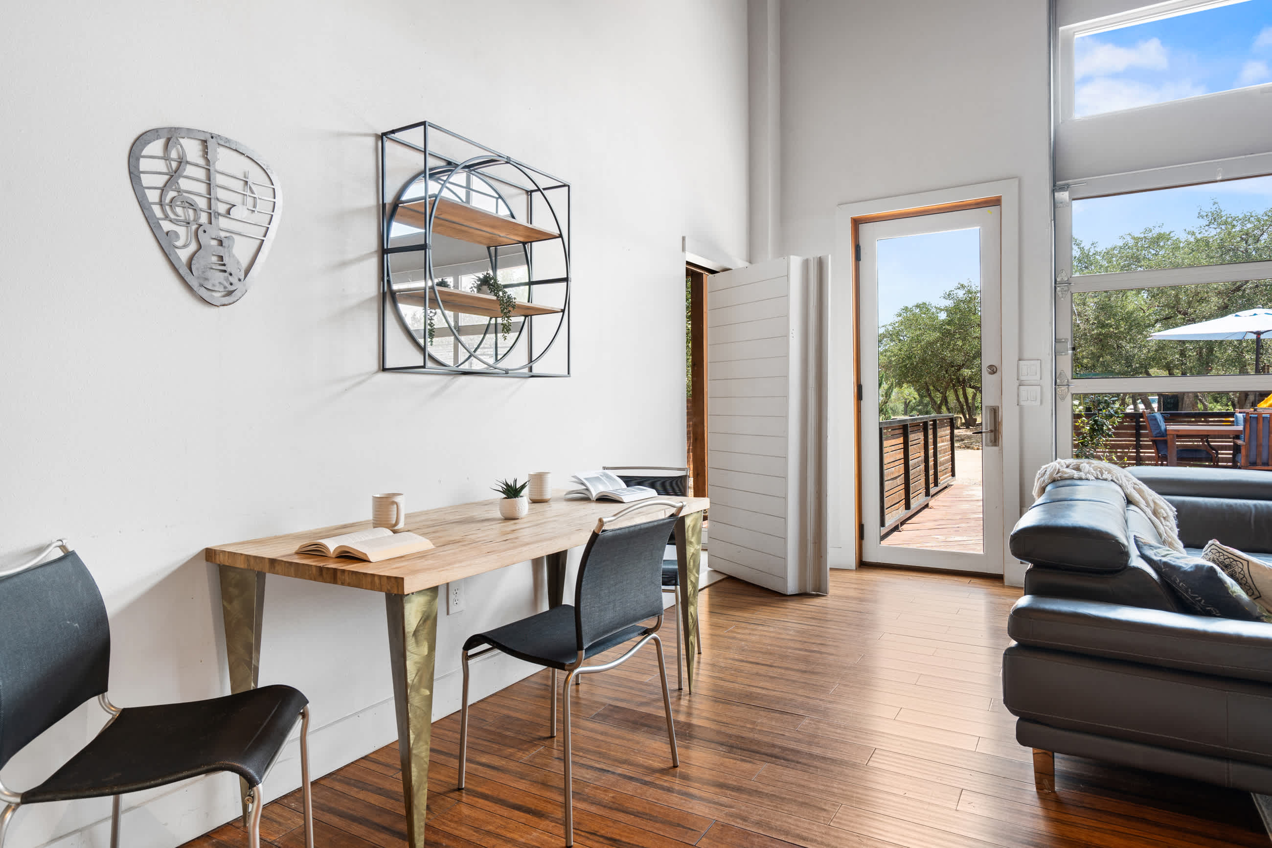 A bright dining area features a light wood table with metal legs and two black chairs, set against a white wall with decorative shelving. A glass door leads to an outdoor deck, offering views of trees.