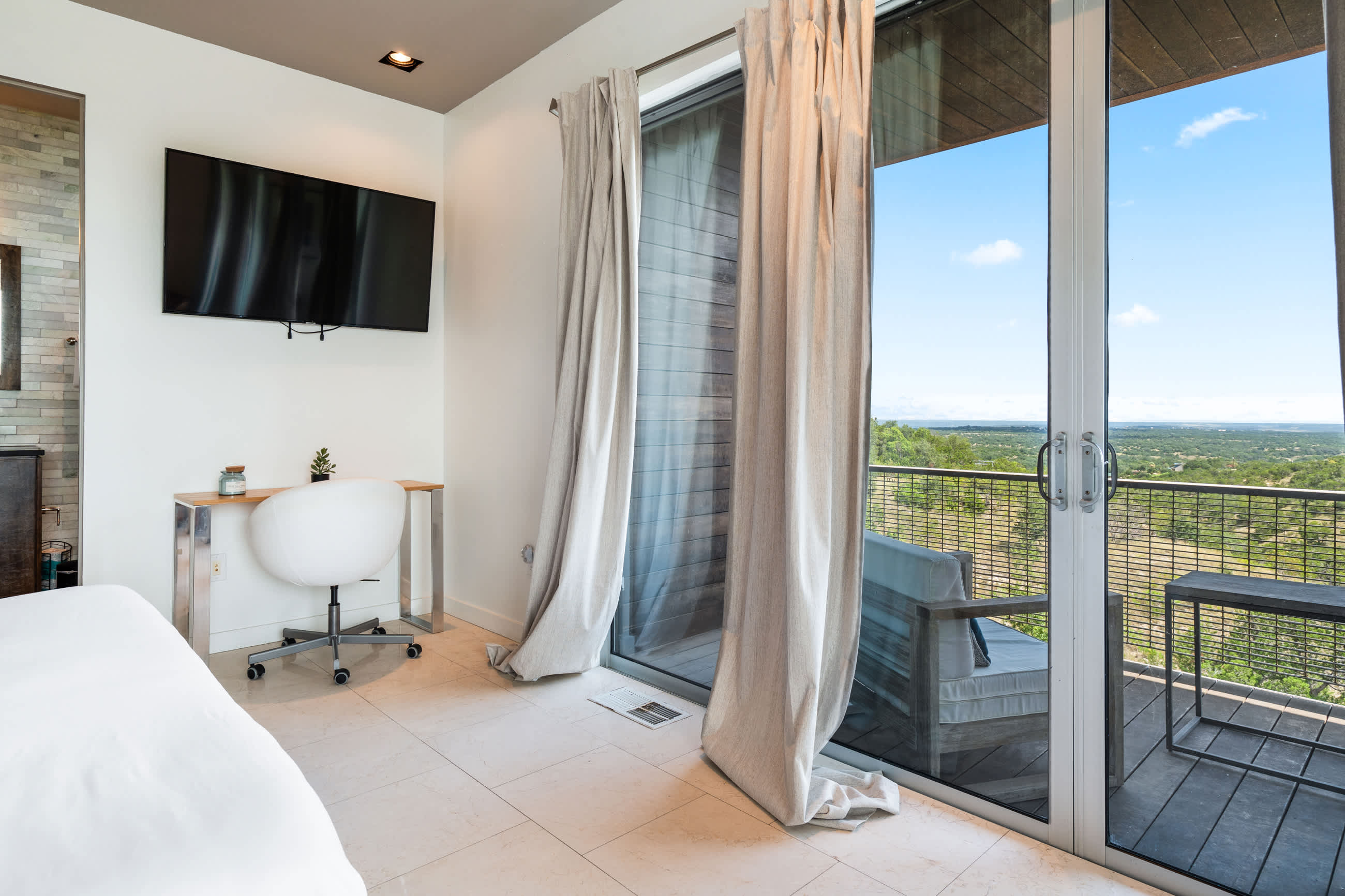 A corner of a modern bedroom featuring a white desk and chair, a wall-mounted TV, and sliding glass doors leading to a private balcony with outdoor seating and a distant view.