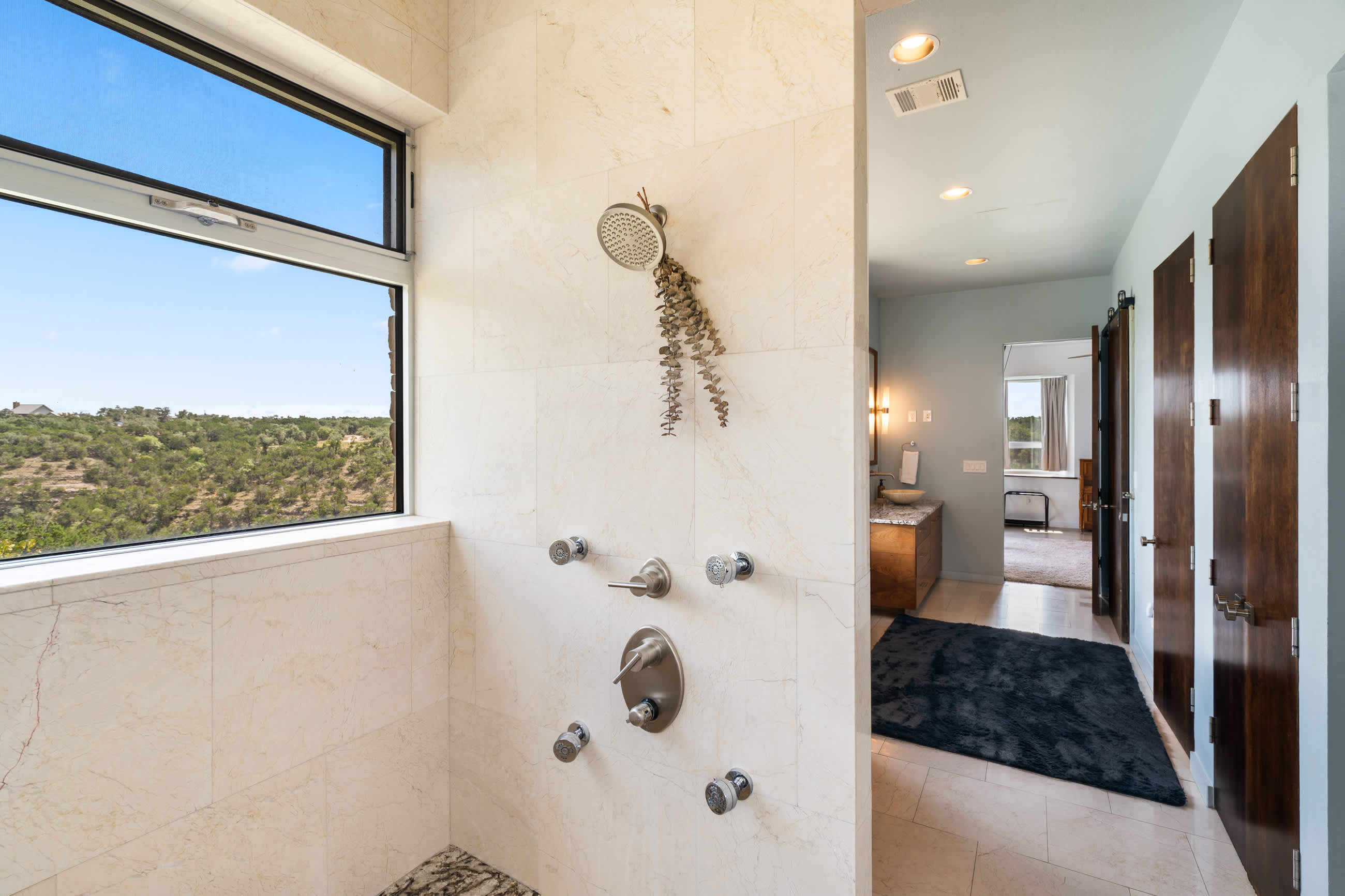 Modern walk-in shower with light-colored tile walls and multiple shower heads, including a rainfall shower. A large window provides natural light and a view of the outdoors.