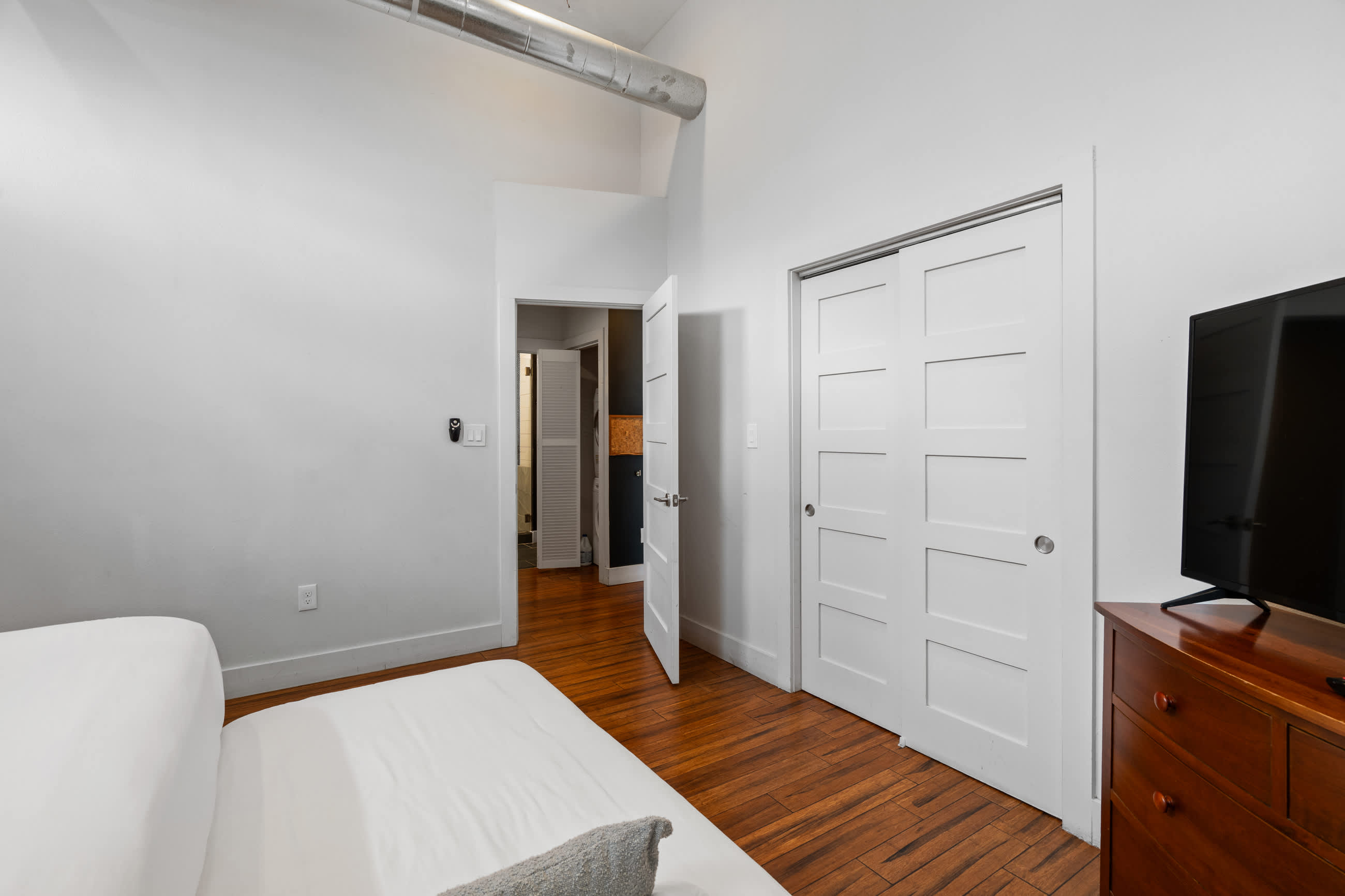 Another angle of the bedroom, highlighting the dark hardwood floors and white walls. Two white paneled doors are visible, one open to reveal a glimpse of a hallway, and the other a closet.