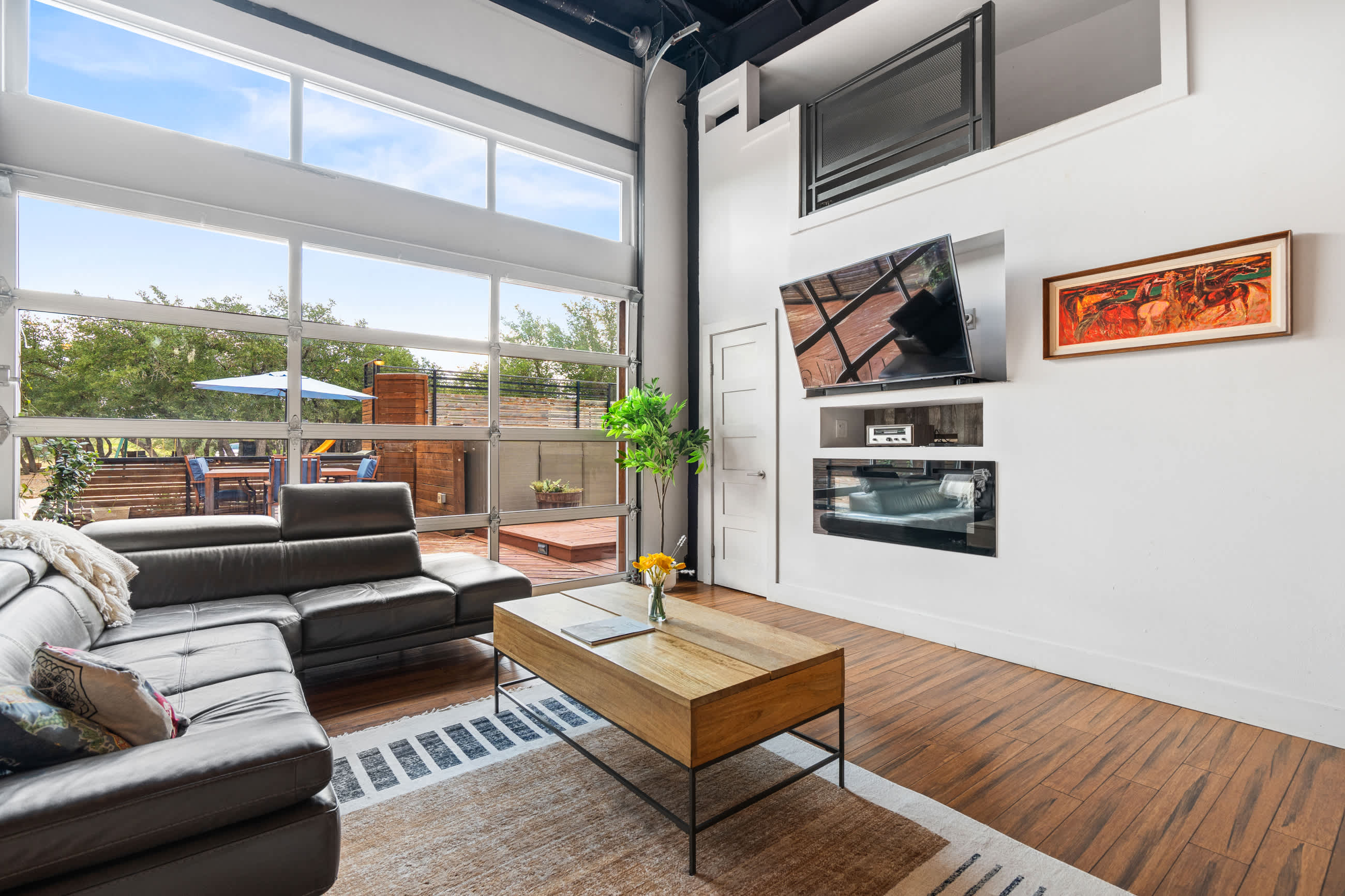 A modern living room features a large black sectional sofa, a wooden coffee table, and rich hardwood floors, with a wall-mounted TV above a sleek fireplace. A full-height glass garage door opens to an outdoor deck.