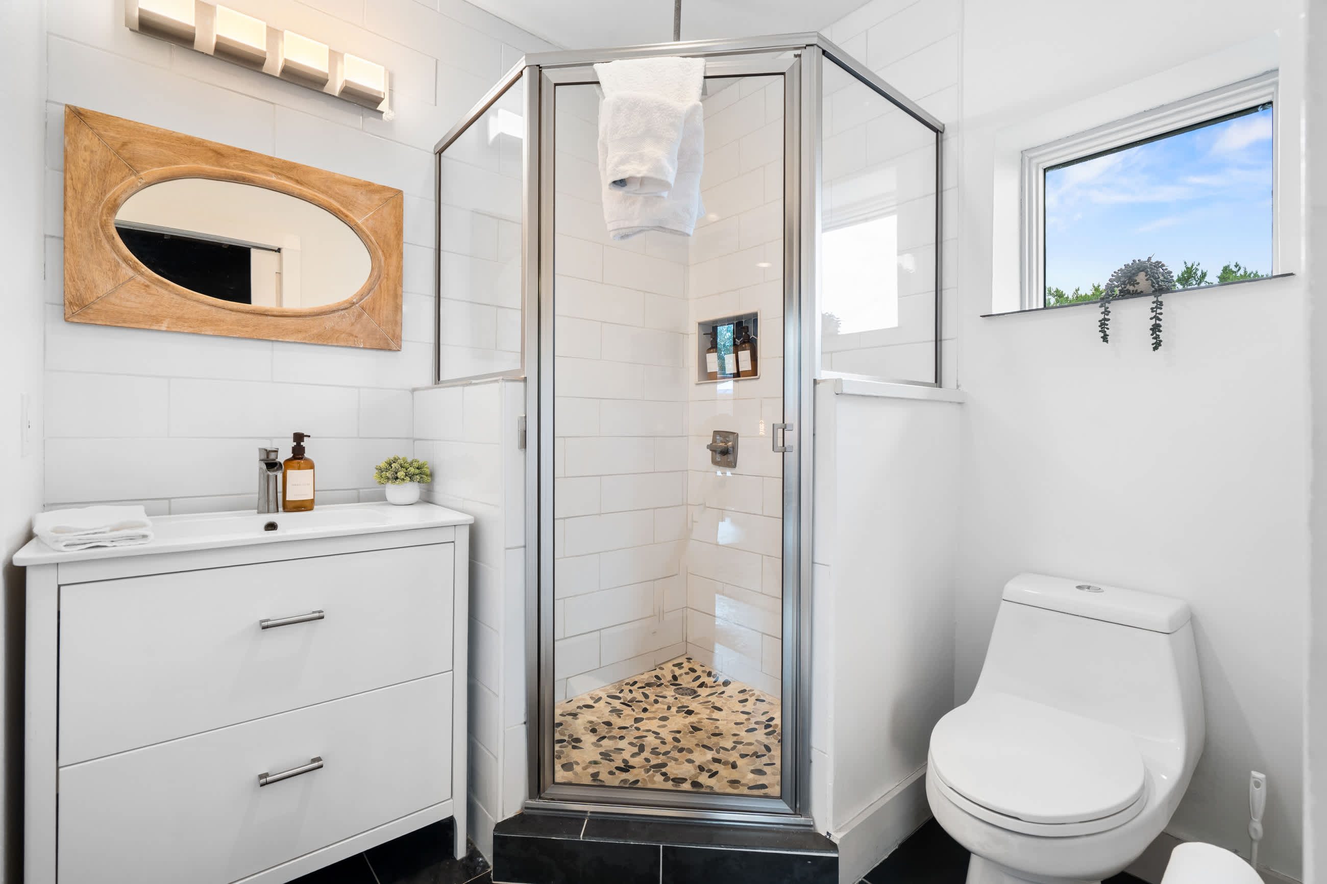 Modern bathroom with a glass-enclosed corner shower featuring a pebble floor and white subway tiles. A white vanity with a sink and a toilet are also visible, along with a small window.
