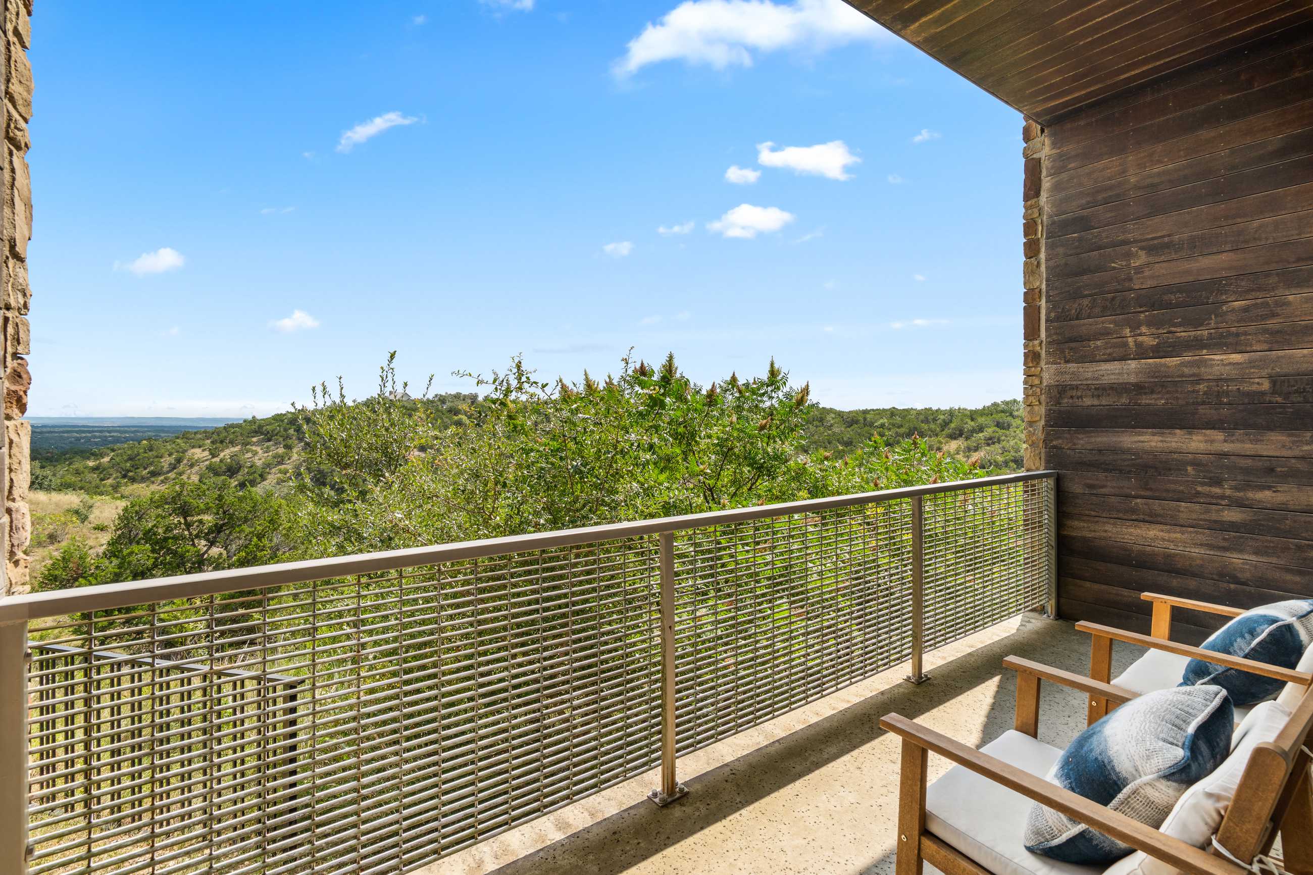 Balcony with a metal grid railing, offering expansive views of rolling green hills and a bright blue sky with scattered clouds. Two wooden chairs with white cushions and blue patterned pillows are visible.