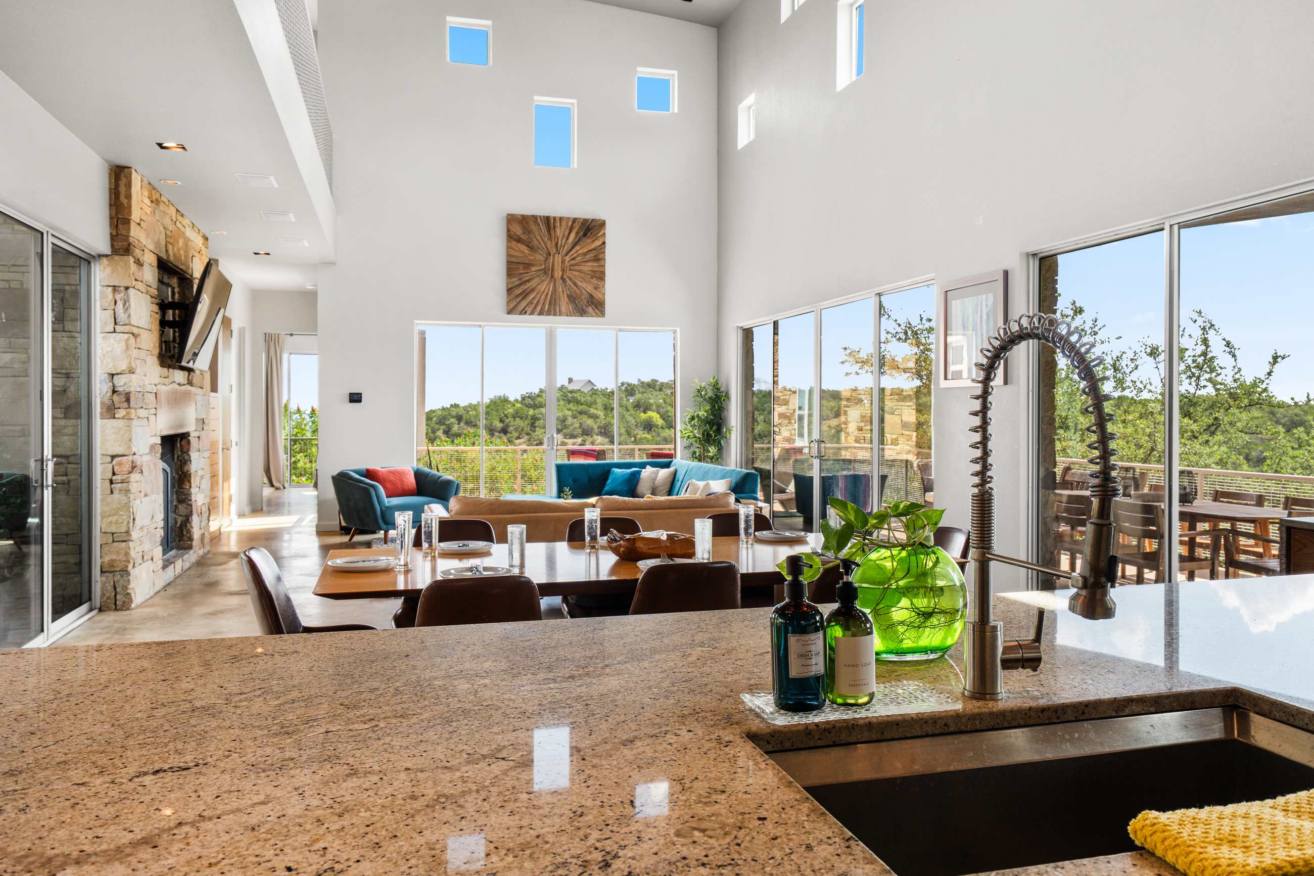 An expansive open-concept living space viewed from the kitchen island, featuring a comfortable seating area with blue sofas and a large stone fireplace. Floor-to-ceiling windows offer panoramic views of the hilly landscape, and a wooden art piece adorns the high wall.