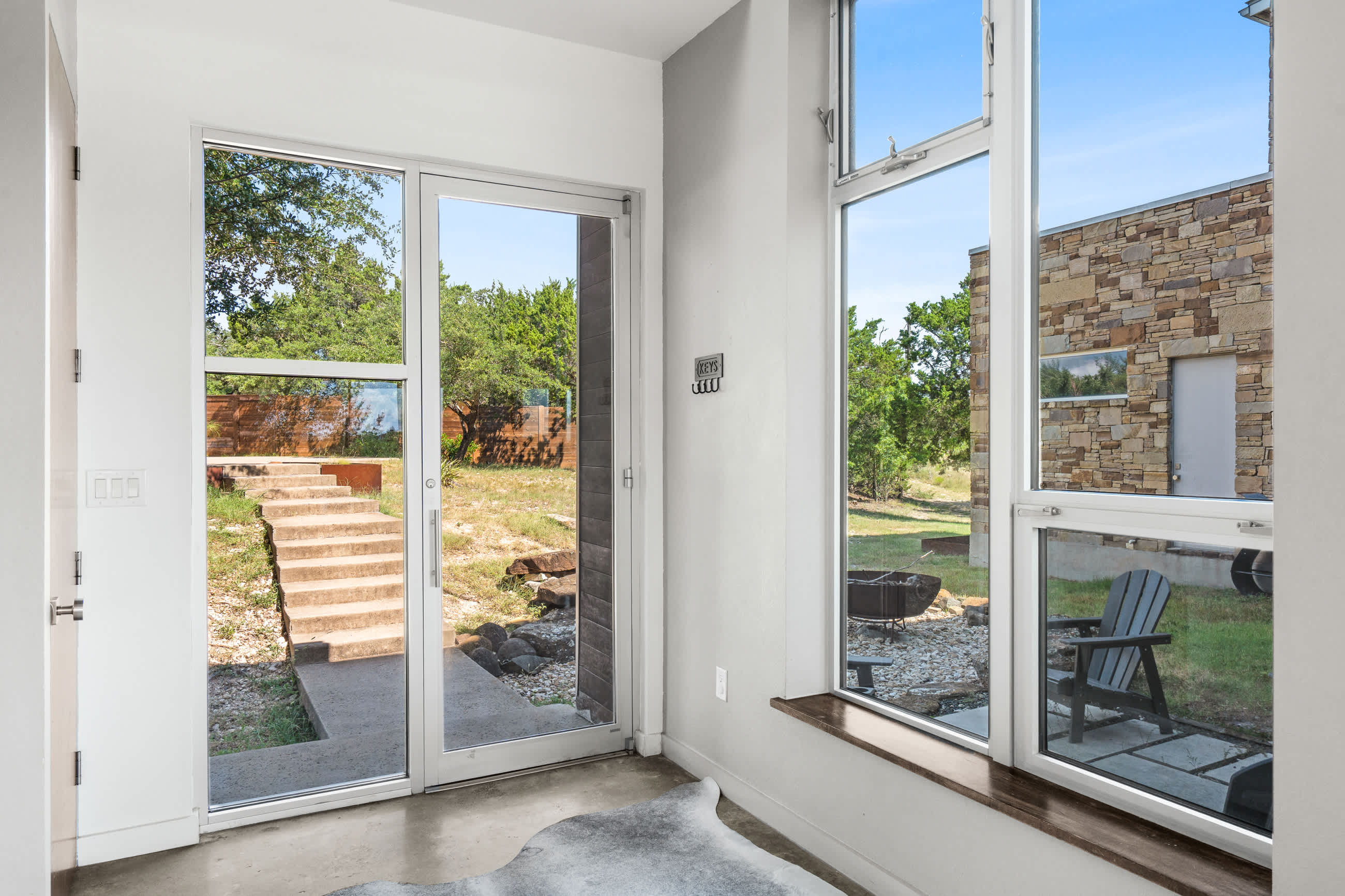 A bright foyer with polished concrete floors and a cowhide rug, featuring a glass front door and large windows offering views of the outdoor patio and backyard.