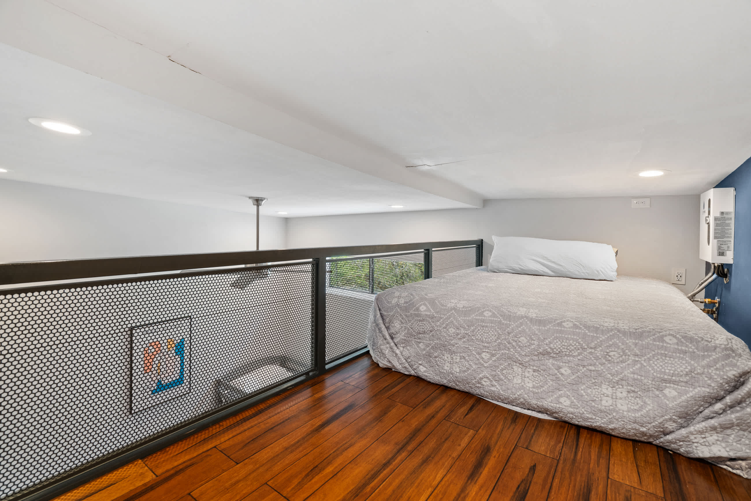 Another view of the loft bedroom, showing the queen bed, dark hardwood floors, and the metal mesh railing. The low ceiling and a white shiplap wall are prominent features.