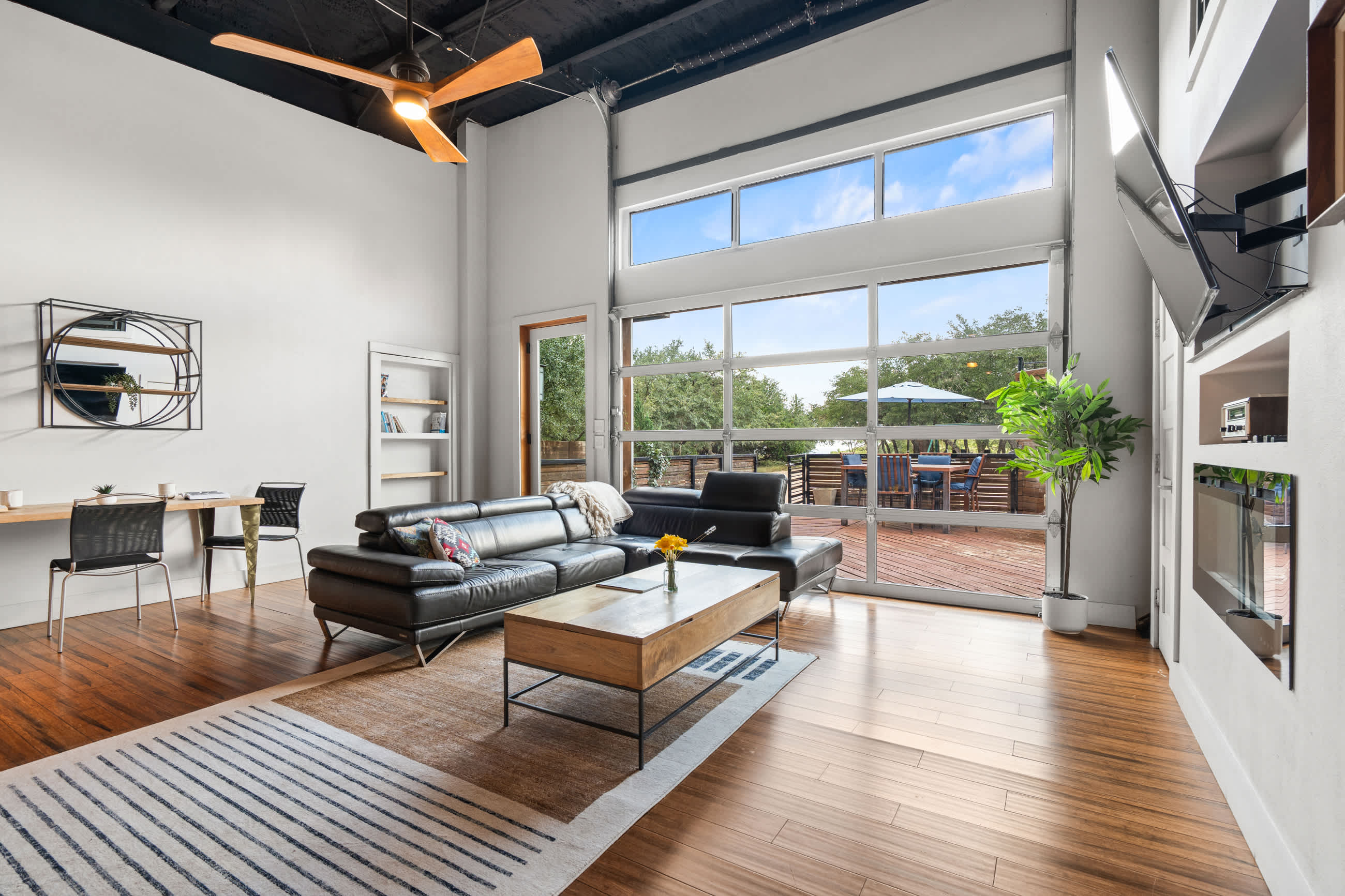 An open-concept living area features a black sectional sofa, a wooden coffee table, and a dining nook with a round mirror, all on hardwood floors. A large glass garage door offers views of the outdoor space, and a ceiling fan hangs overhead.