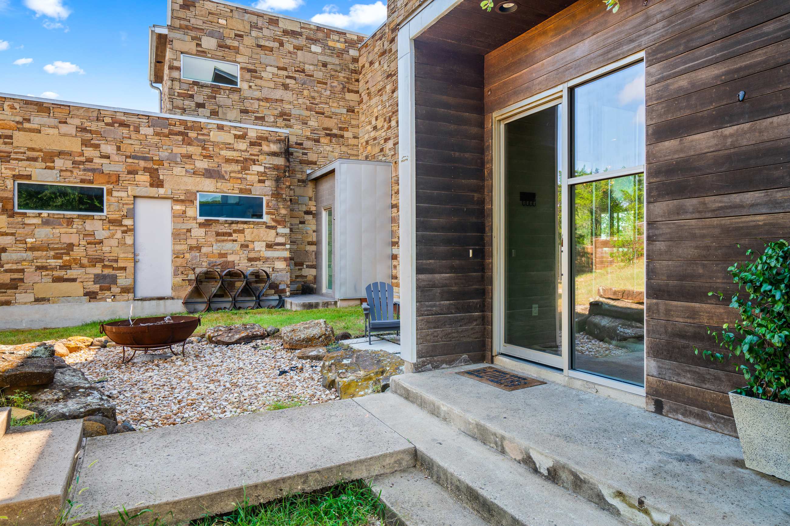 A side view of the house exterior featuring a concrete walkway leading to a glass door, a fire pit area with gravel and large rocks, and a mix of stone and wooden siding.