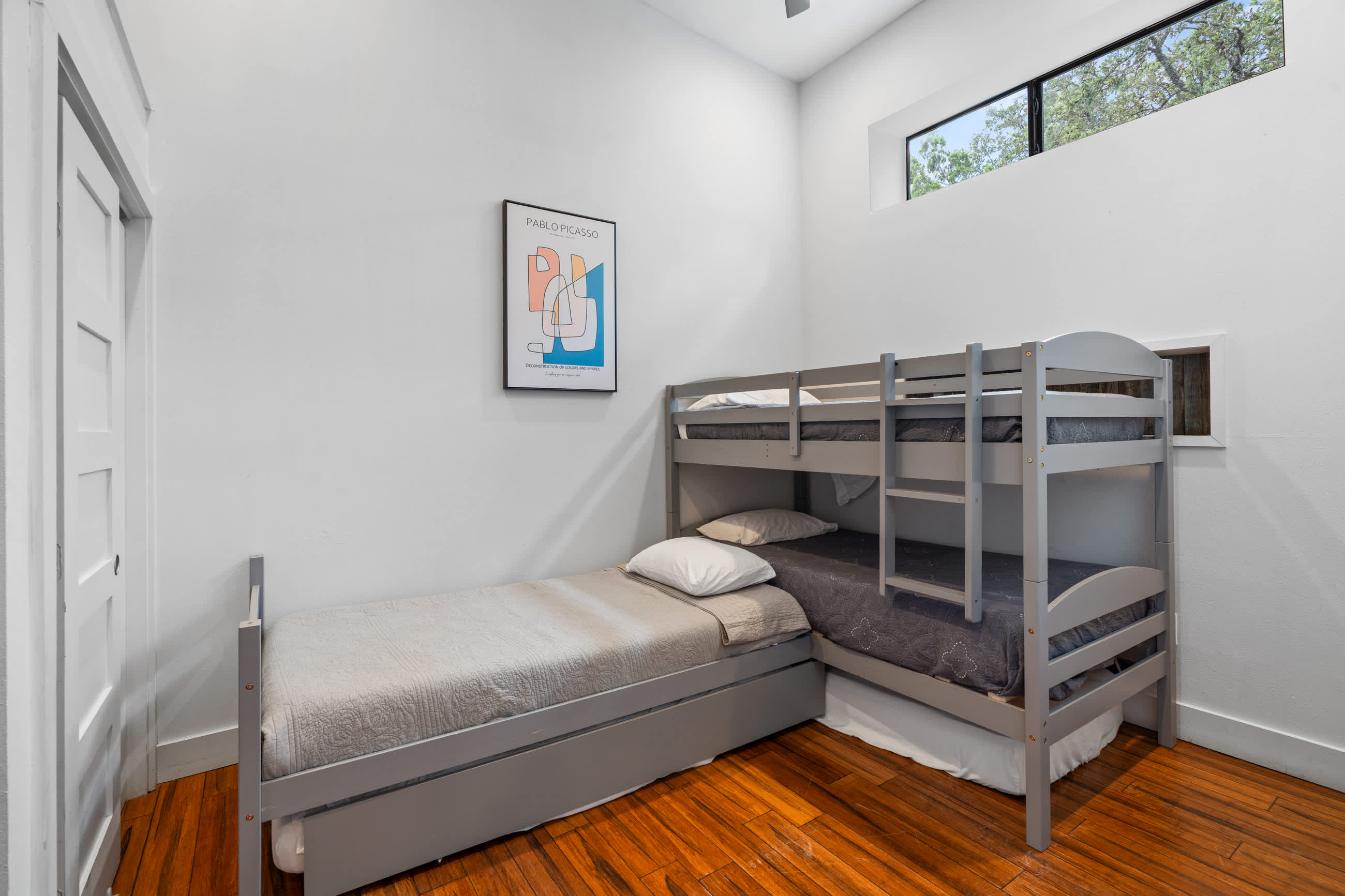 Kids' bedroom featuring a gray bunk bed and a separate gray twin bed with a trundle, all on dark hardwood floors. White walls and a high window are visible, along with a colorful abstract art print.