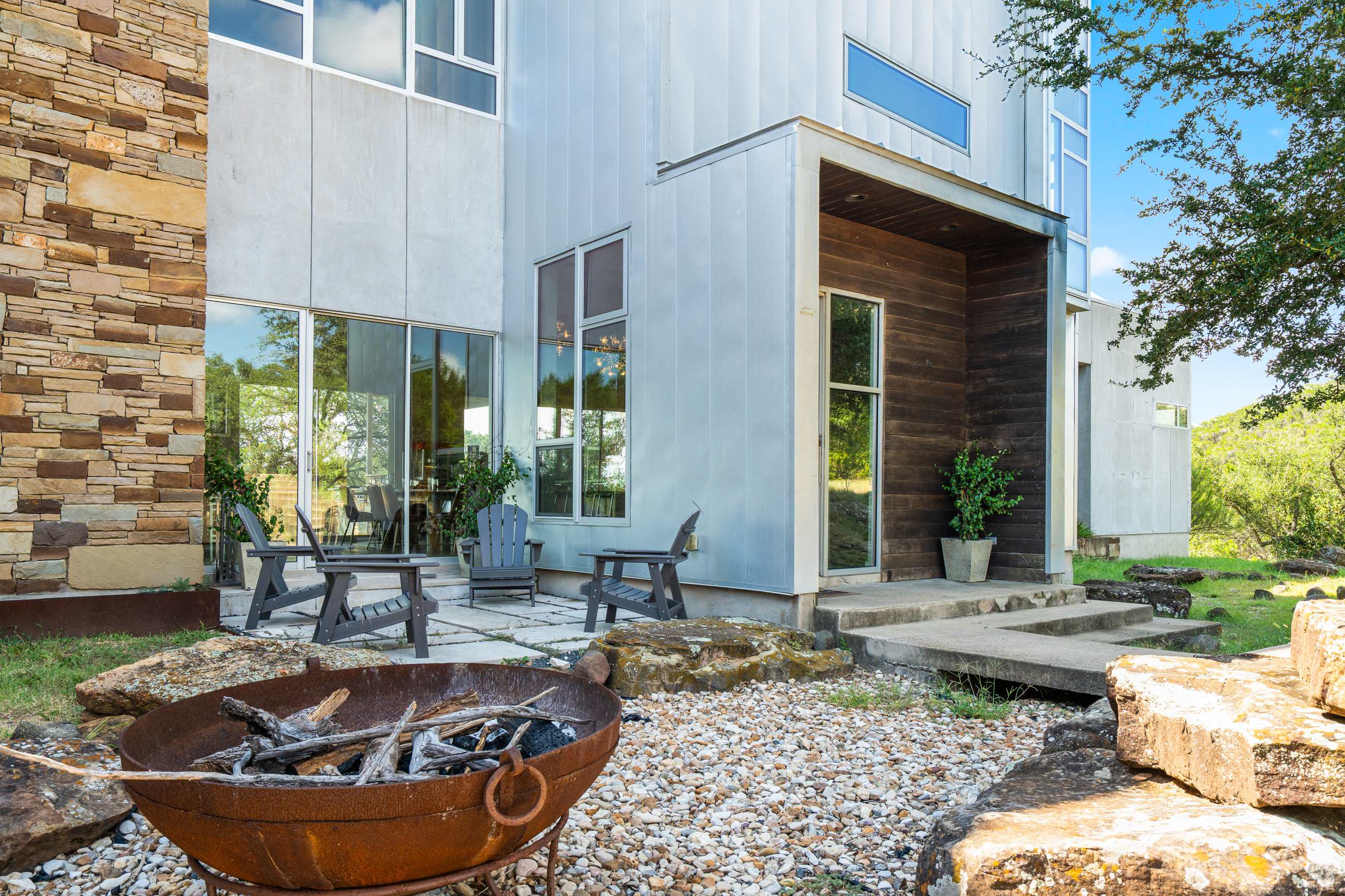 A rustic fire pit with logs sits on a gravel and rock bed in a spacious backyard, with a modern house featuring stone and metallic siding in the background.