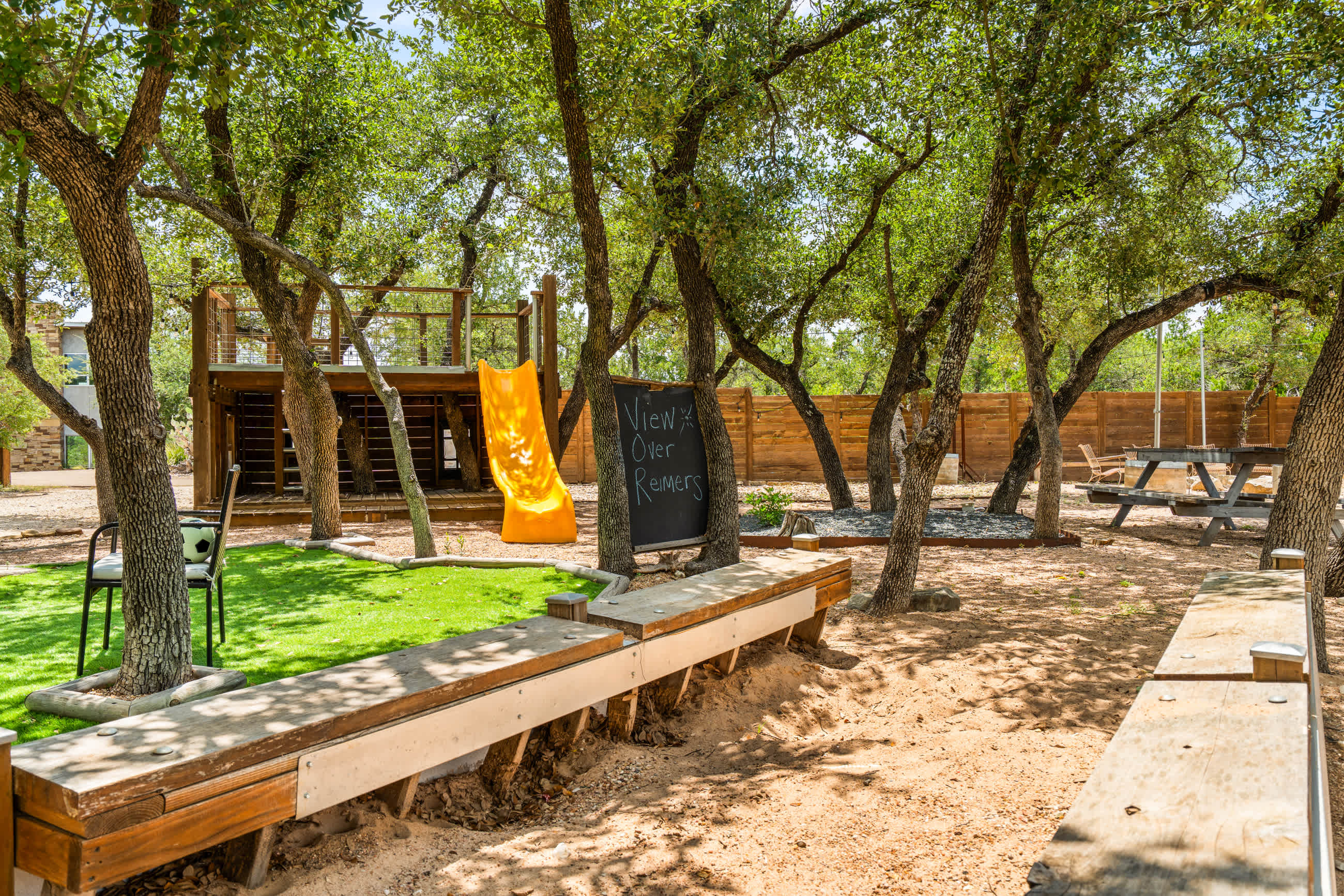 Expansive outdoor play area with a wooden play structure and orange slide, an artificial turf patch, and a long wooden bench. Mature trees provide ample shade over the gravel ground.