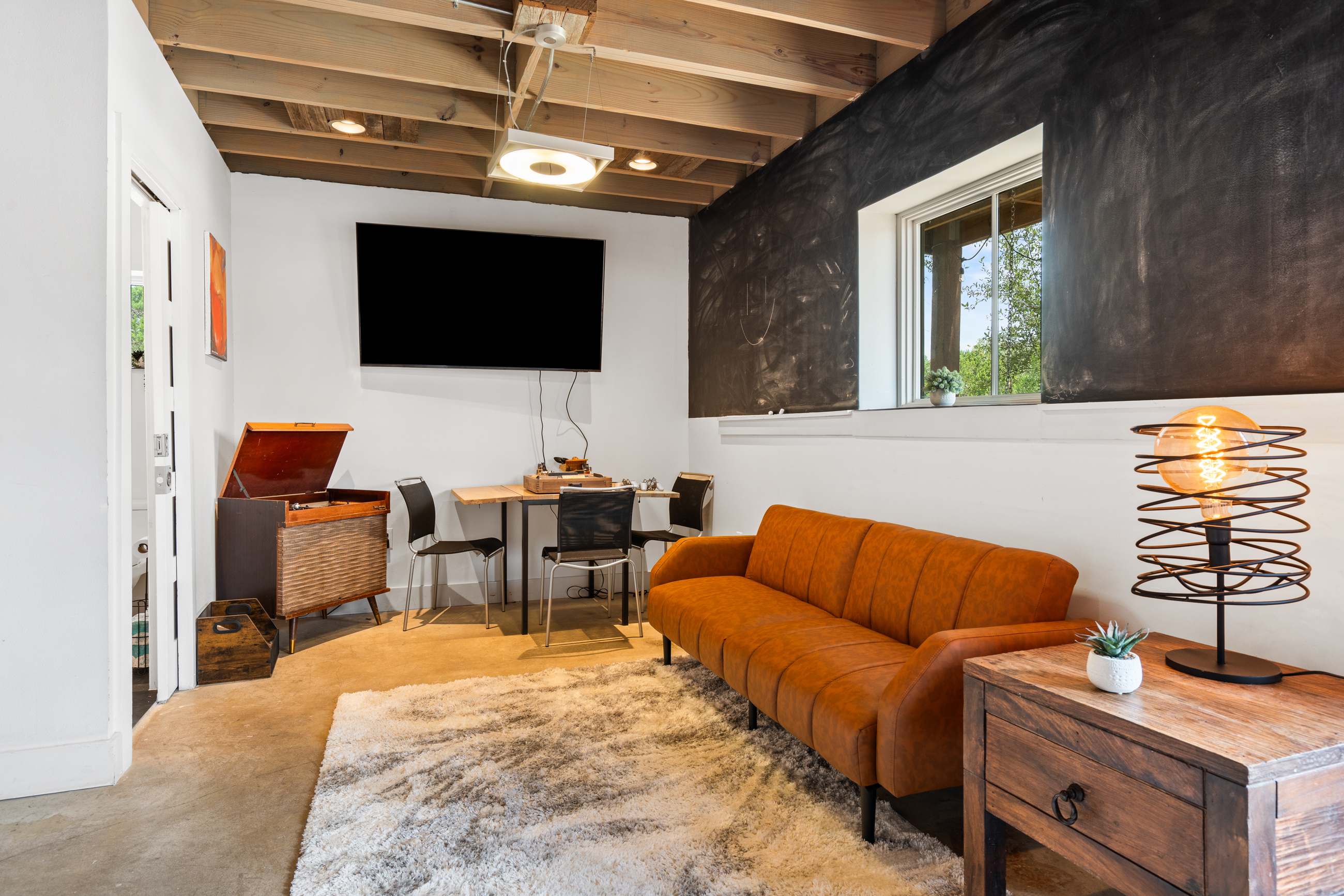 A cozy recreation room features an orange sofa, a large wall-mounted TV, and a vintage record player cabinet. The room has an exposed wooden beam ceiling, concrete floors, and a chalkboard wall with a window.