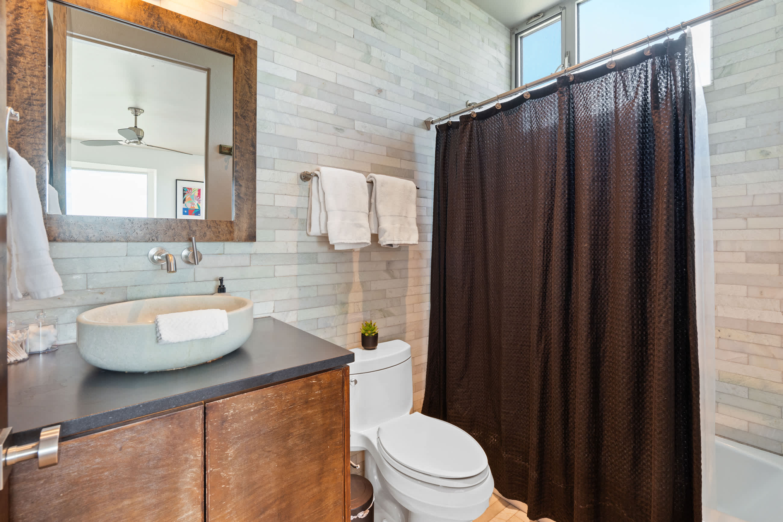 Bathroom with a unique stone vessel sink on a dark countertop and a wooden vanity, featuring a dark brown shower curtain and white subway tile walls. A toilet is visible next to the shower.