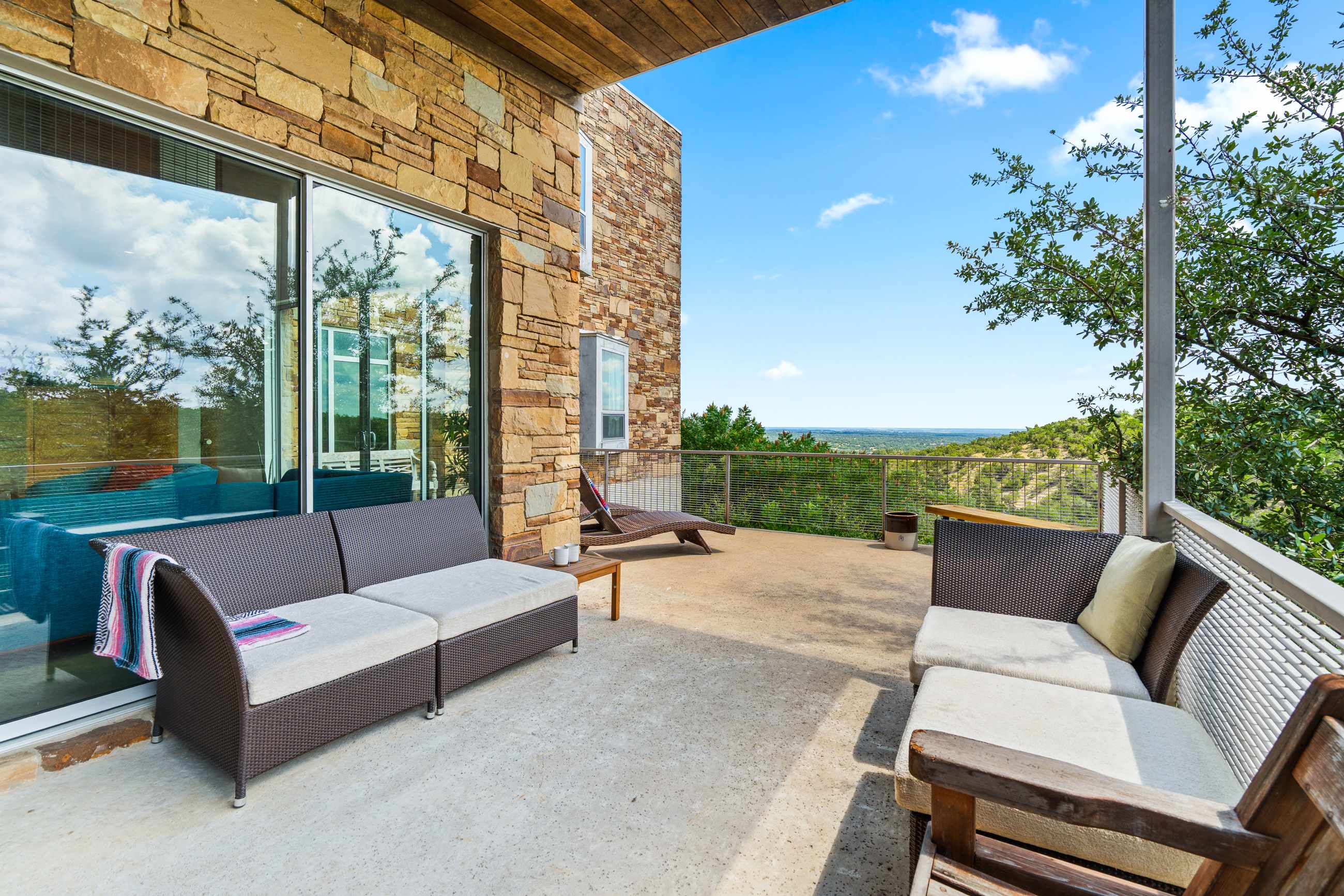 A covered outdoor patio features contemporary wicker lounge sofas with cushions and a wooden bench, facing a panoramic vista of rolling hills and a distant body of water. The adjacent building wall is clad in natural stone.