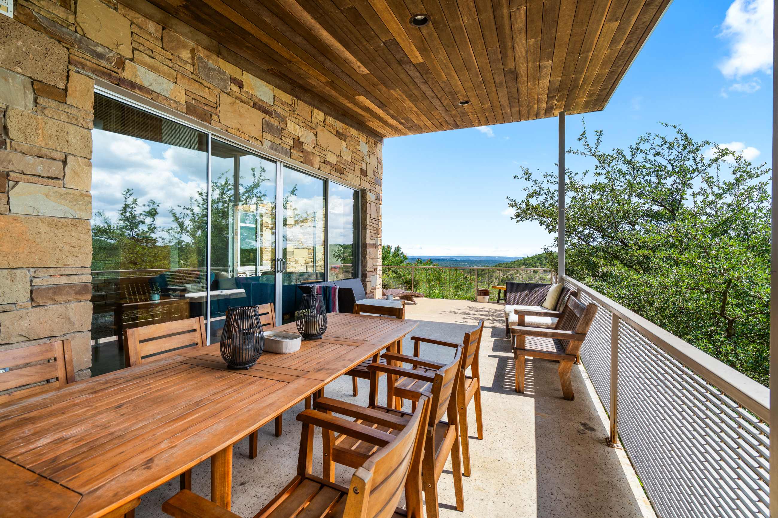 A spacious covered outdoor patio features a long wooden dining table with chairs, alongside a lounge area, all set against a stunning panoramic view of rolling hills and a distant lake under a blue sky. The ceiling is wood-paneled, and the walls are stone.