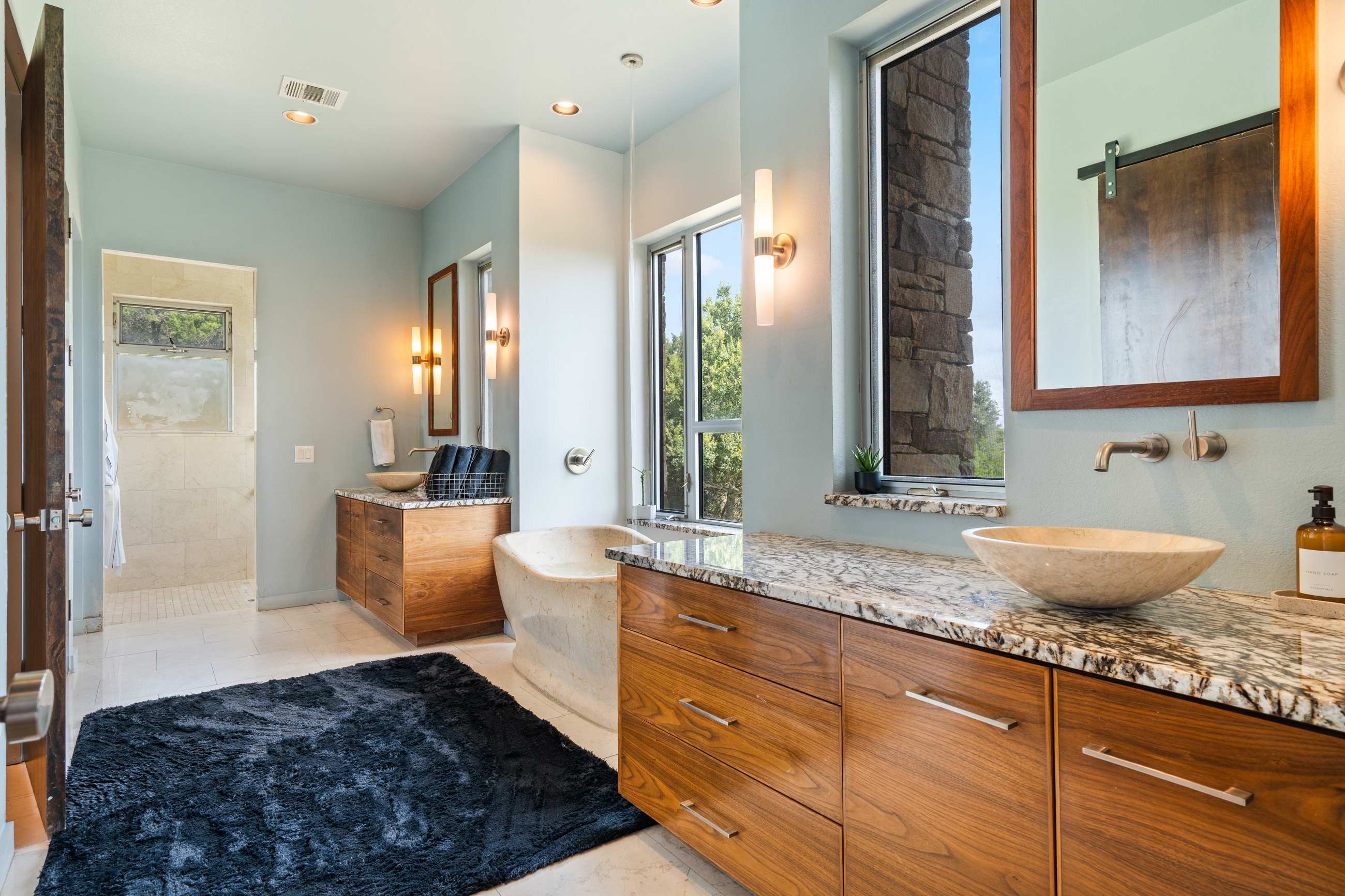 Luxurious bathroom with light blue walls, two wooden vanities with granite countertops and vessel sinks, and a freestanding stone bathtub. A large window offers a view of the outdoors.