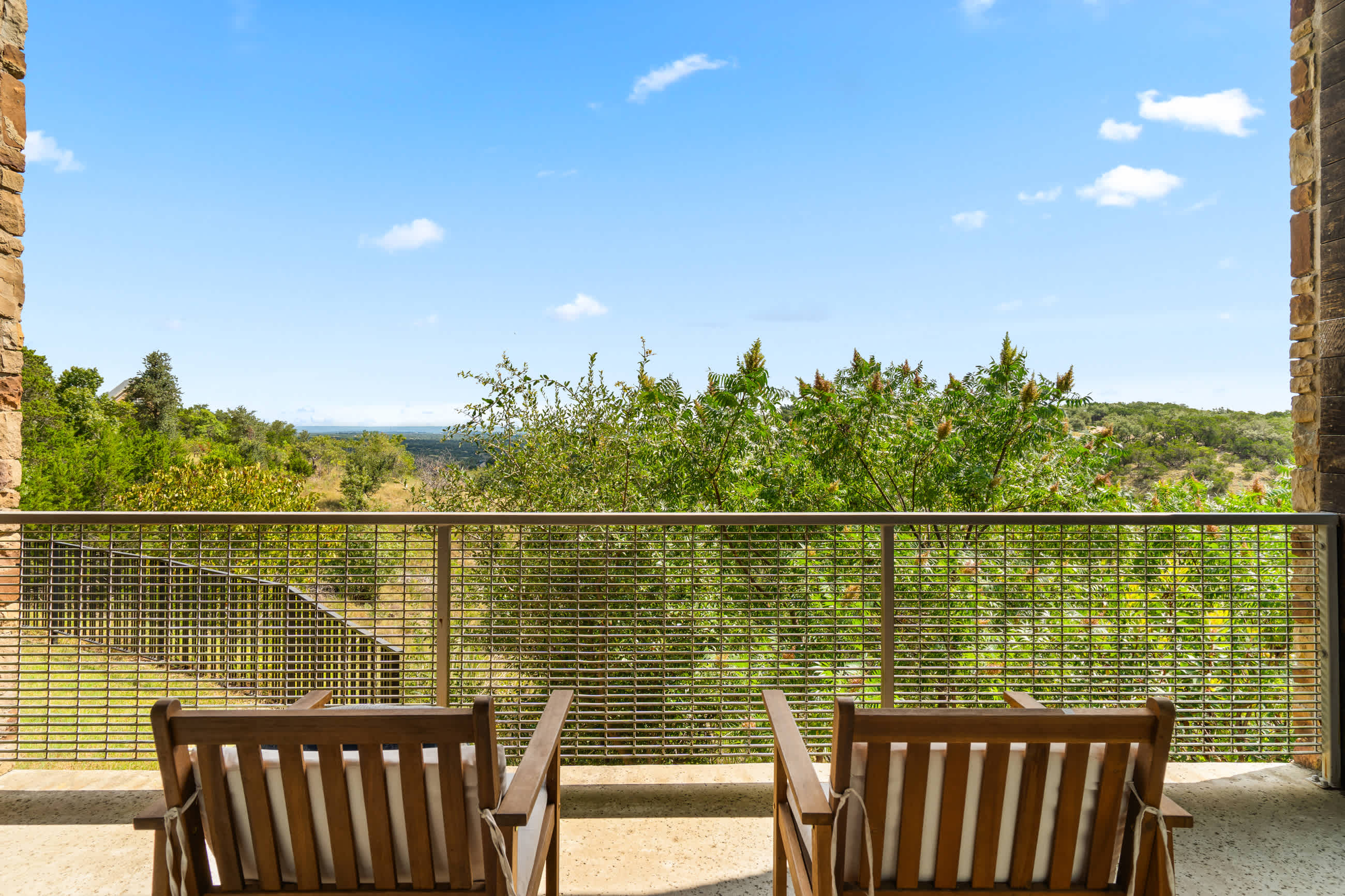 Scenic balcony view from behind two wooden chairs with white cushions, showcasing a vast landscape of green hills under a clear blue sky with white clouds. The metal grid railing provides an unobstructed vista.
