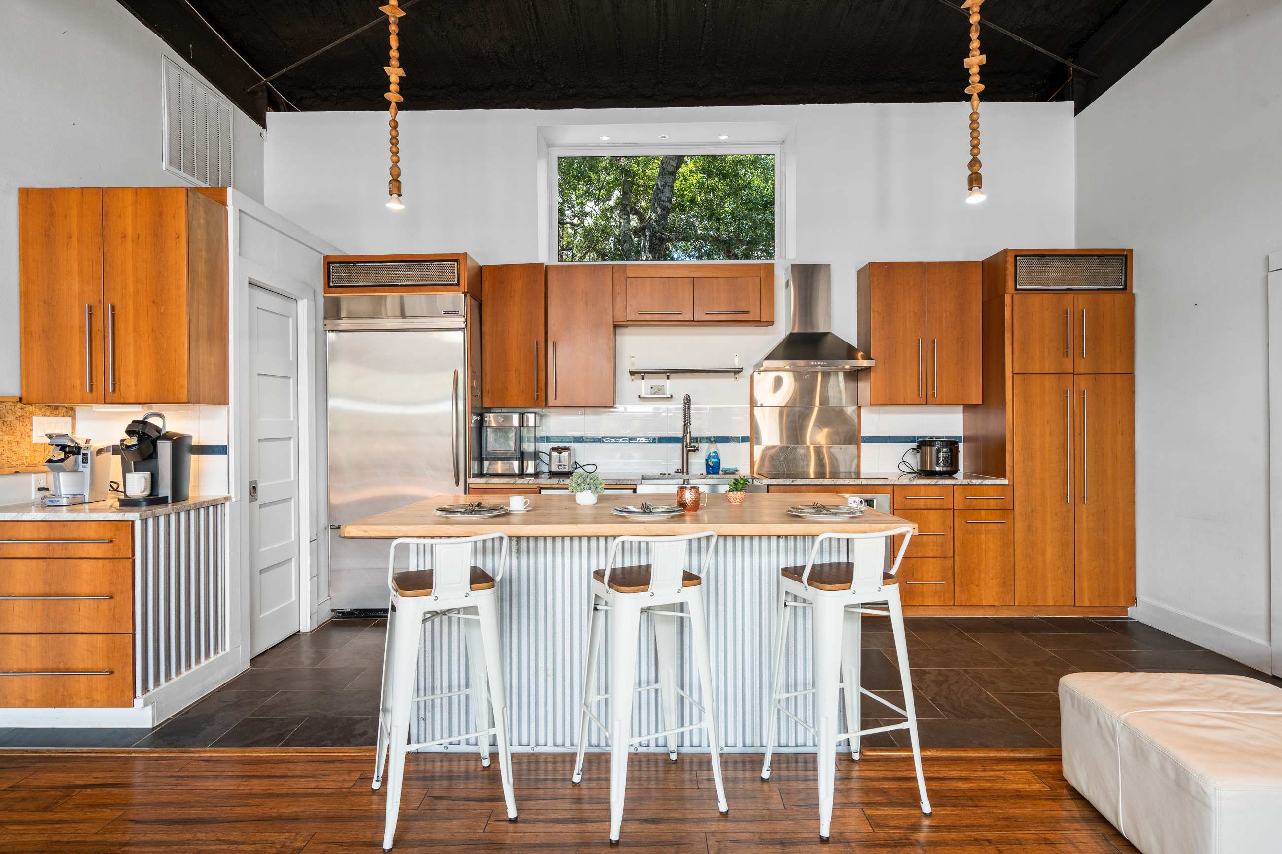 A modern kitchen features light wood cabinetry, stainless steel appliances, and a large island with a butcher block countertop and corrugated metal base. Three white bar stools are pulled up to the island, and a large window overlooks greenery.
