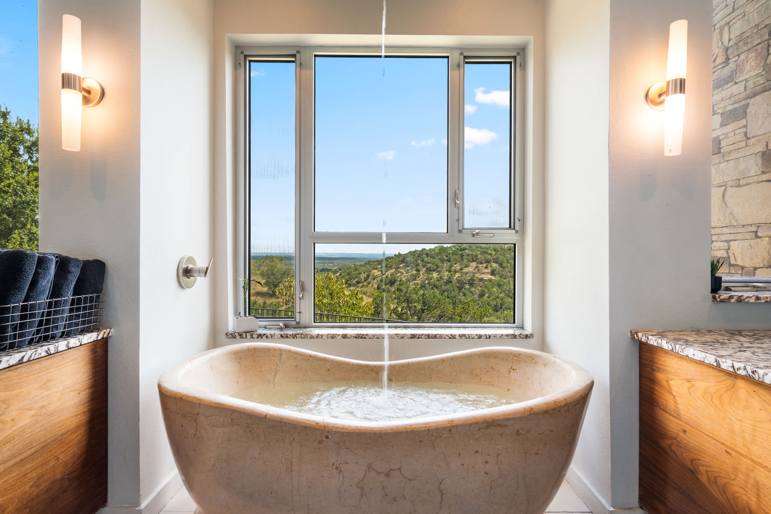 Close-up of a unique, light-colored stone freestanding bathtub with water filling from a ceiling-mounted faucet, set against a large window offering a scenic view of the landscape.
