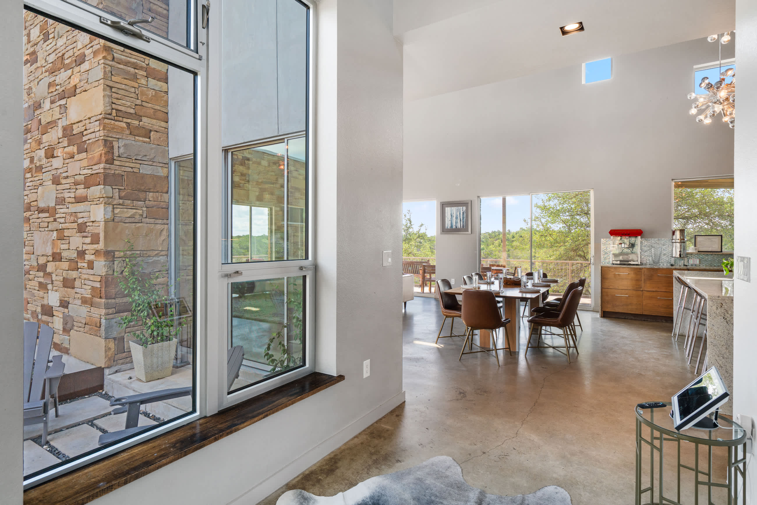 A view from the foyer into an open-concept dining area with a large wooden table and leather chairs, and a modern kitchen beyond, all brightly lit by large windows.