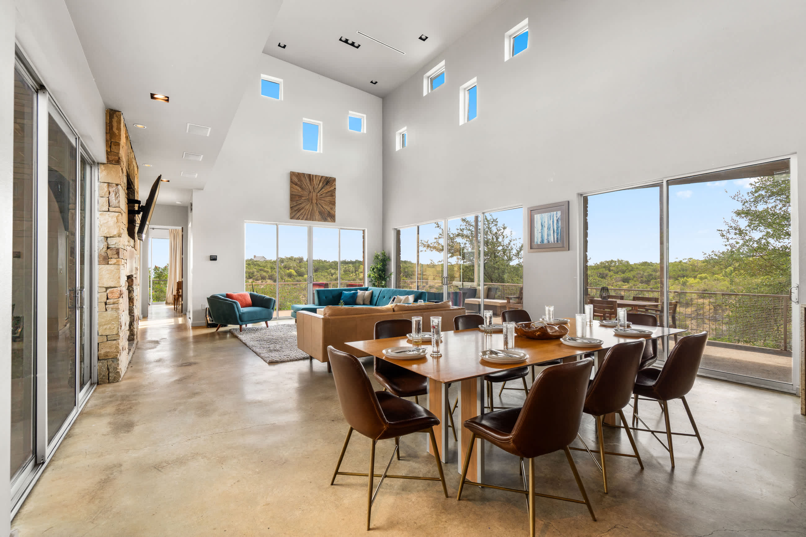 Open-concept dining area with a long wooden table and six brown leather chairs, featuring high ceilings, large windows with outdoor views, and a stone fireplace, adjacent to a living space with a brown sectional sofa.