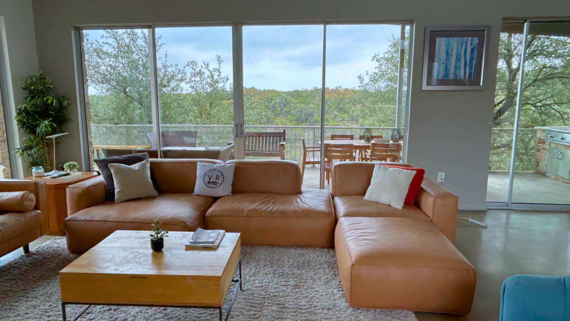 Spacious living room with a large brown leather sectional sofa and a matching smaller sofa, a wooden coffee table, and a light shag rug, facing expansive windows that overlook a wooded outdoor area.