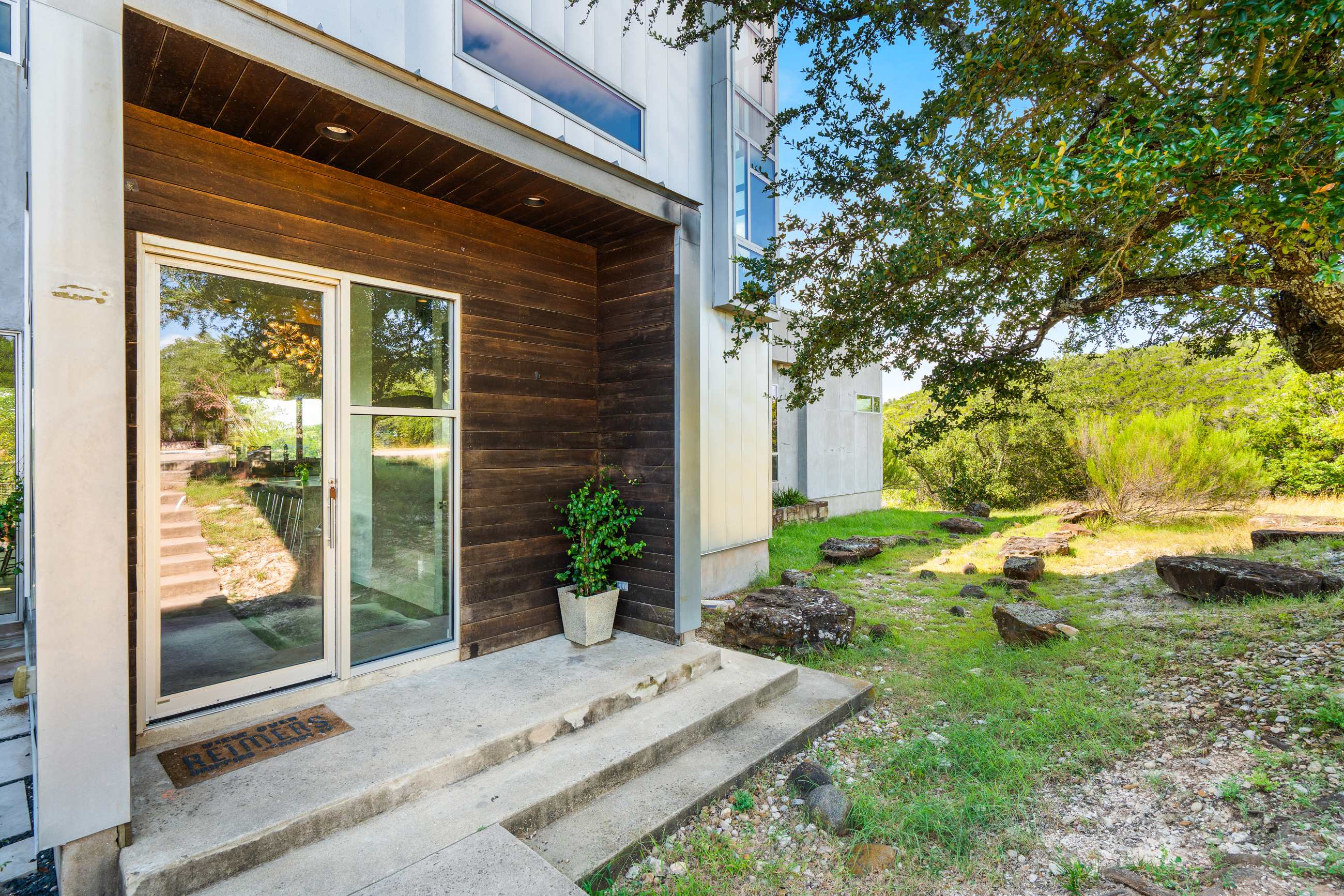 The modern front entrance features a recessed wooden facade with a glass door, concrete steps, and a small potted plant, surrounded by natural landscaping and a large tree.
