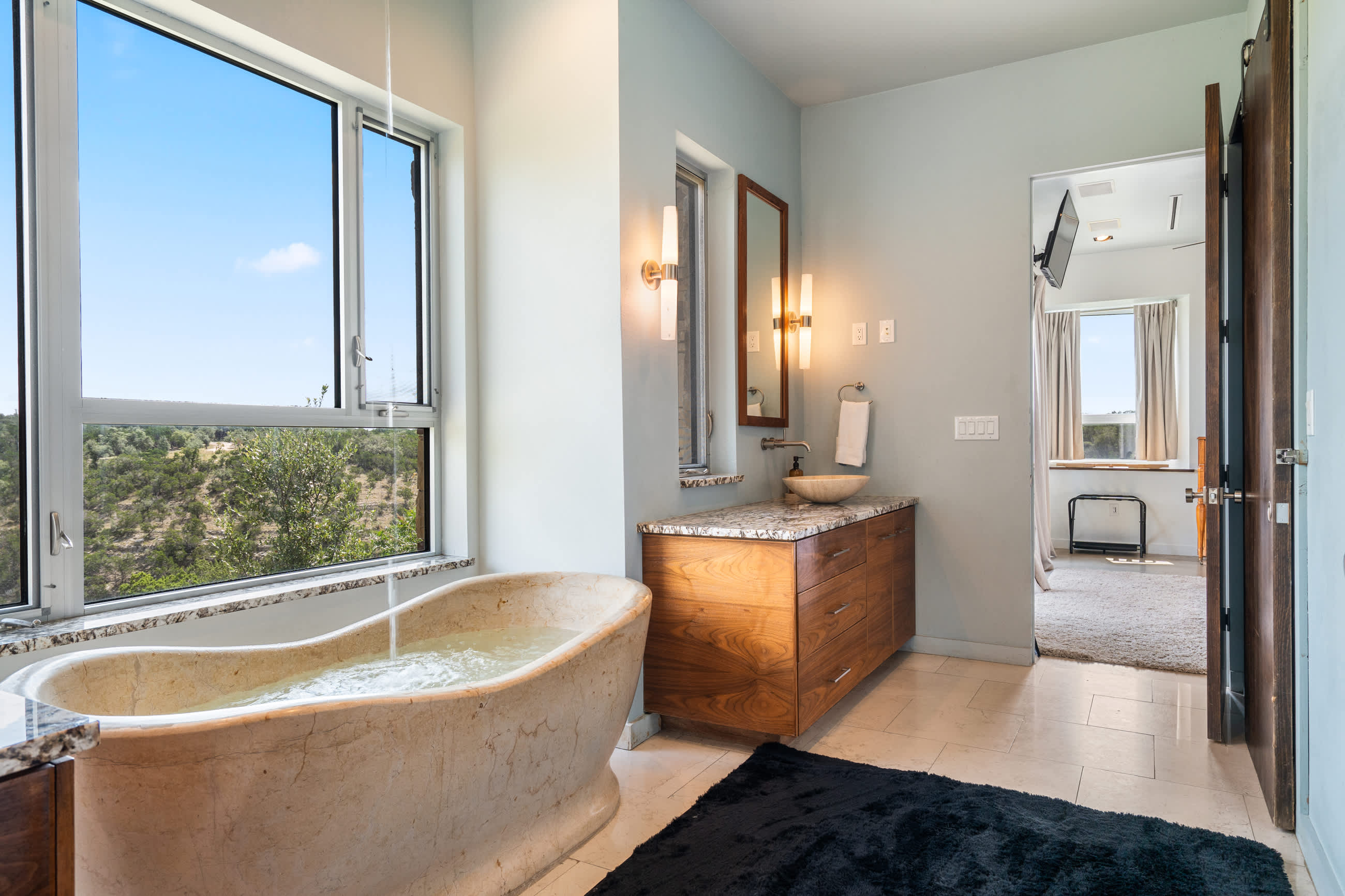 View of a spacious bathroom with a freestanding stone bathtub positioned by a large window overlooking a natural landscape. A wooden vanity with a vessel sink is visible, and a doorway leads to the adjacent bedroom.