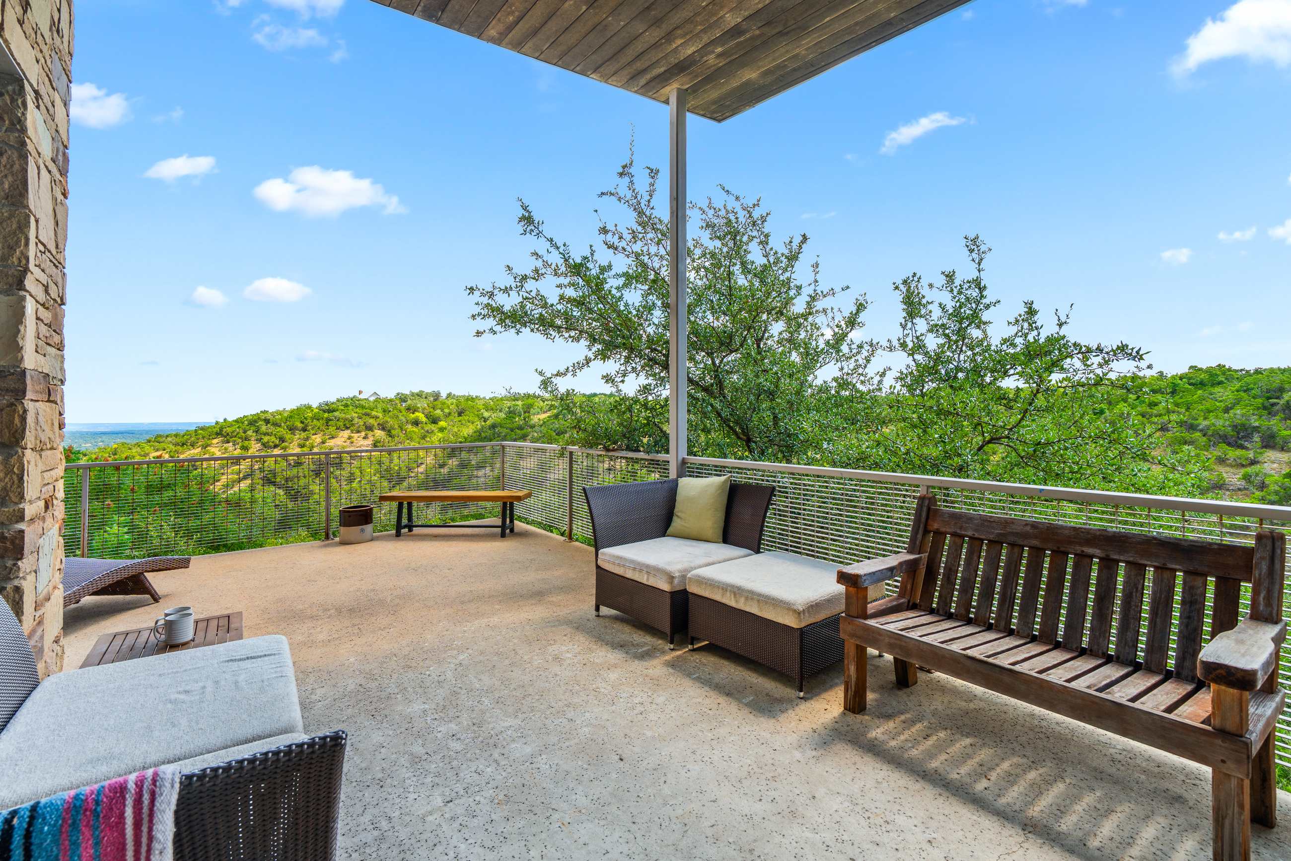 A spacious outdoor patio offers comfortable lounge seating and a wooden bench, providing expansive views of a verdant valley and distant horizon under a bright blue sky. A modern metal railing secures the perimeter.